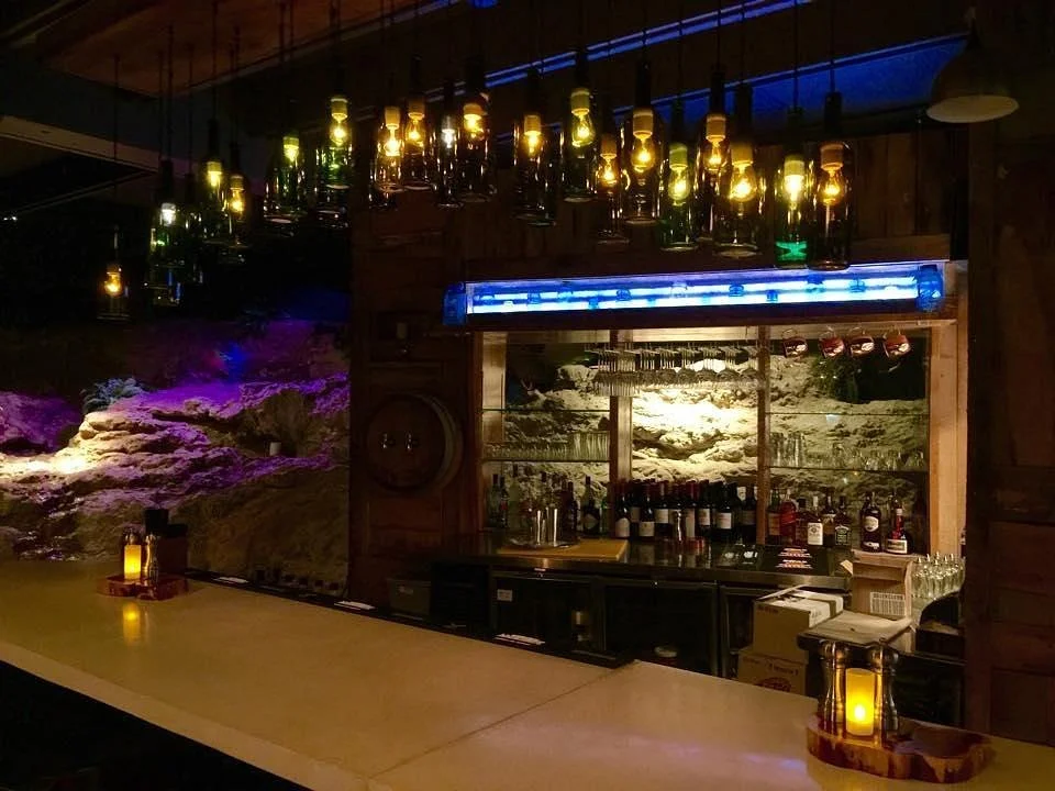 The intimate, warmly lit bar at Grotto Wood-Fired Grill and Wine Cave in Eureka Springs, featuring unique wine bottle chandeliers and a stunning natural rock wall background.