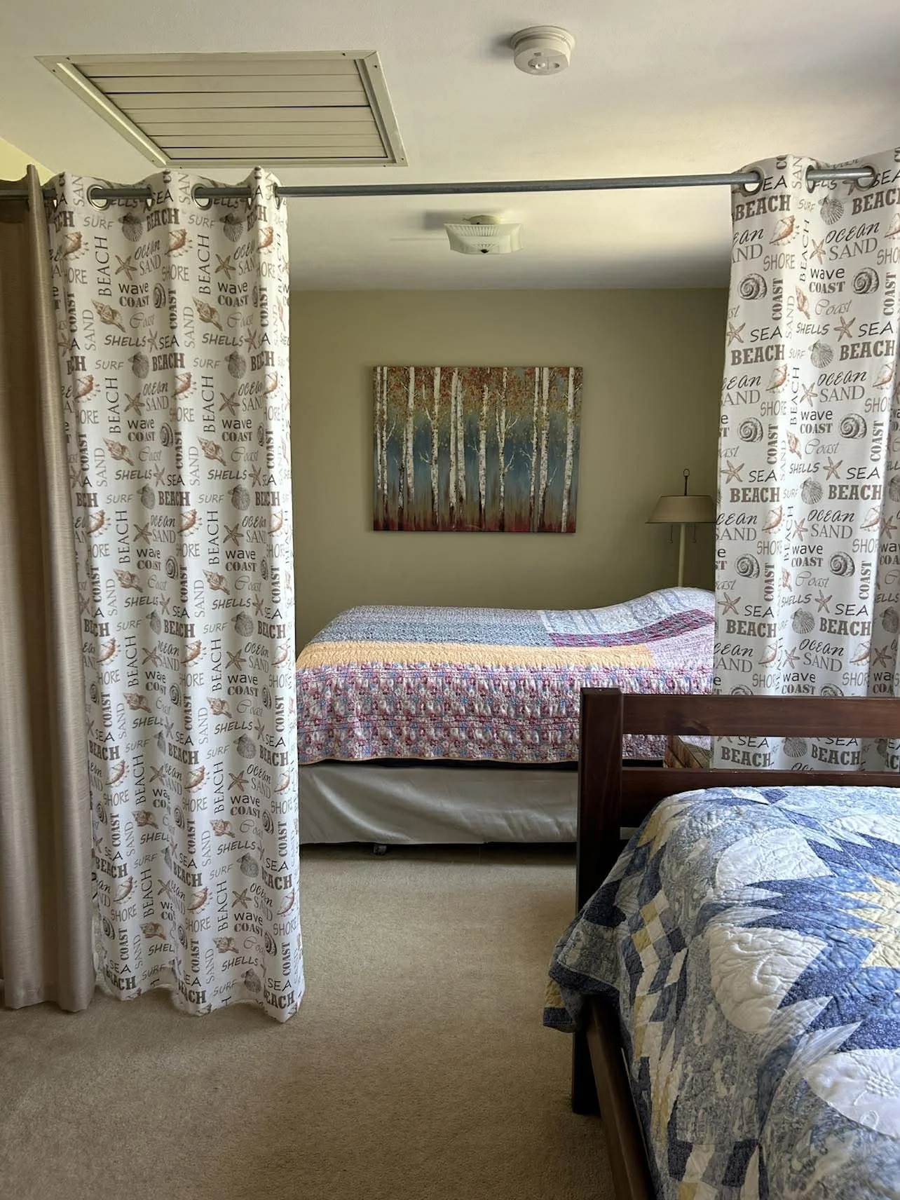 A hotel room with two beds separated by curtains. The bed in the background has a colorful quilt and a painting of trees on the wall above it. The foreground bed has a blue and white quilt. The room has beige carpet, a ceiling vent, and a wall lamp.