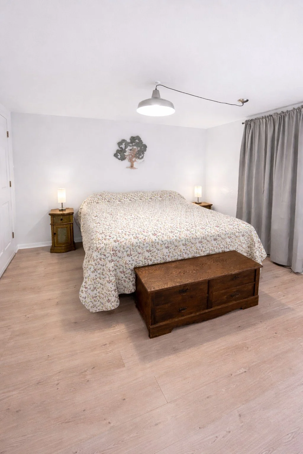 A bedroom with a bed, wooden bench at foot, two nightstands with lamps, a decorative tree on wall, gray curtains, and light wood flooring.