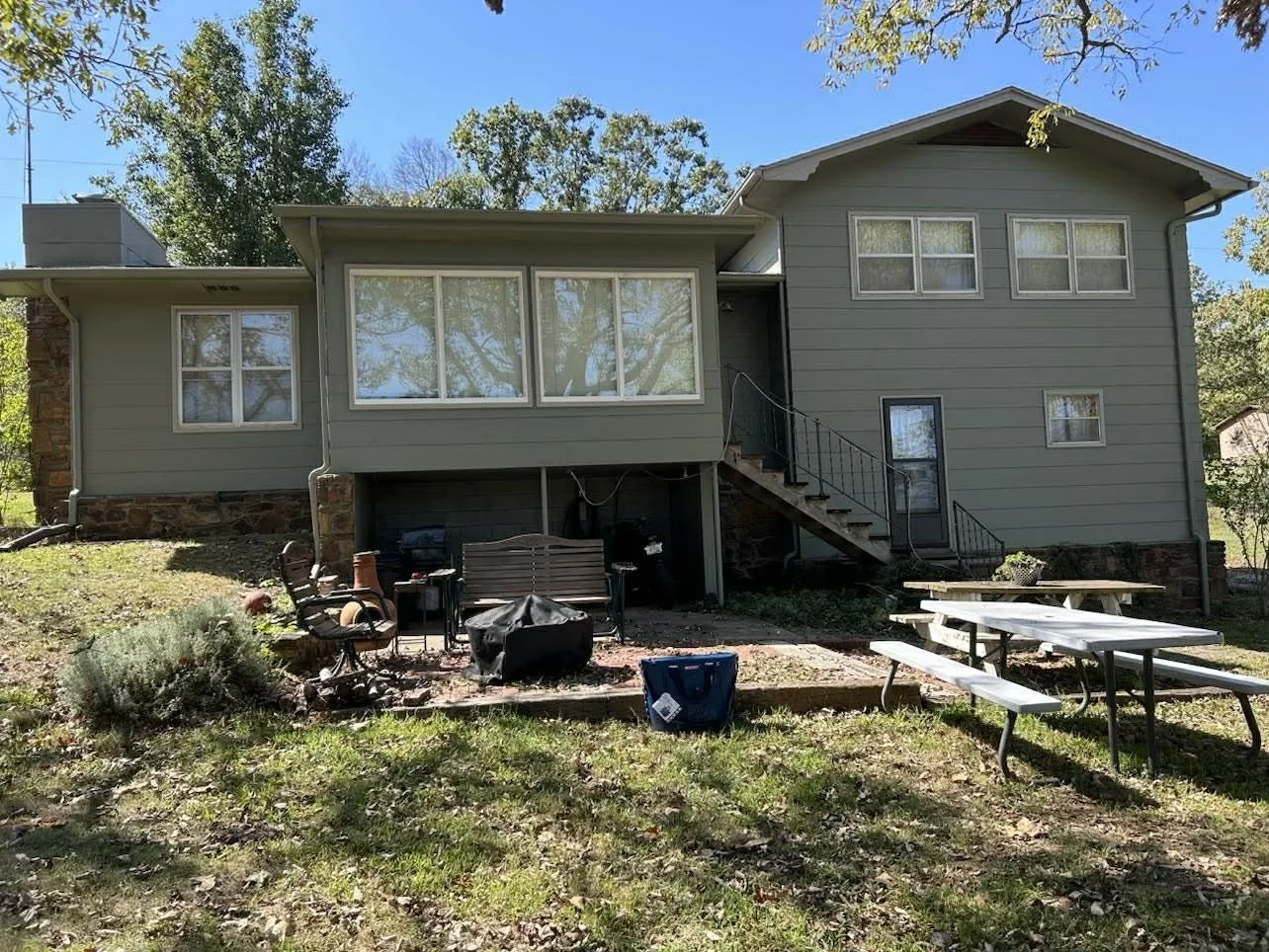 Backyard view of a two-story house called Martha's view on Table Rock Lake, with a staircase leading to a door, outdoor furniture including a bench, chairs, and picnic tables, and a small garden. Trees and clear blue sky in the background.