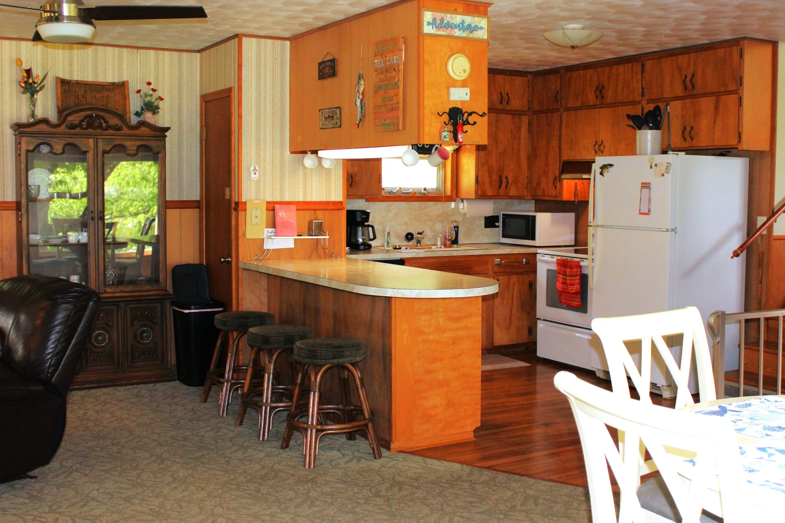 Interior of a vintage kitchen with wooden cabinets, a small window, white refrigerator, microwave, coffee maker, and wall decor including a sign that says 'Adventure'. A round kitchen island with bar stools is also visible, along with a dining table 