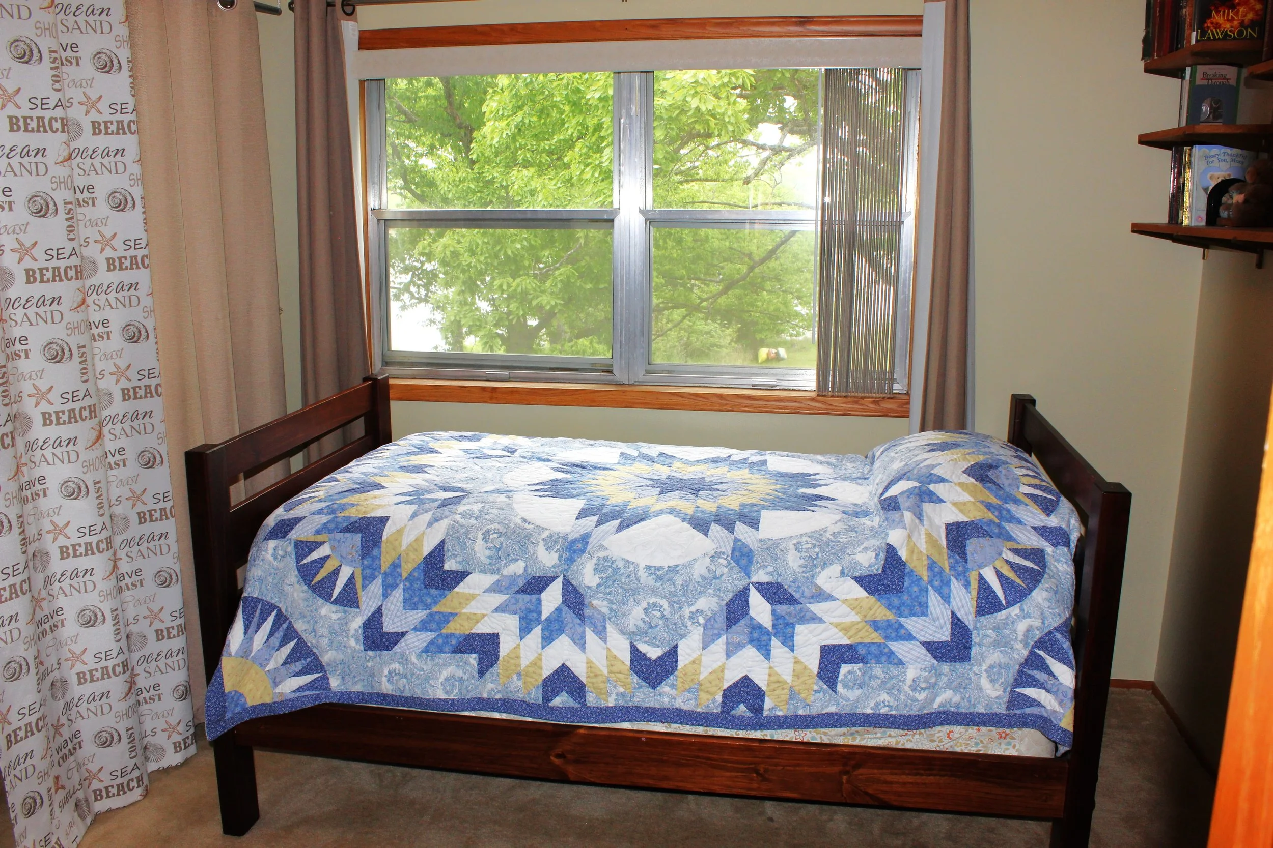 A bedroom with a large window showing green trees outside. There is a bed with a blue, yellow, and white quilt. Beige curtains are on either side of the window, and there are wooden shelves with books on the right side of the image. The floor is carp