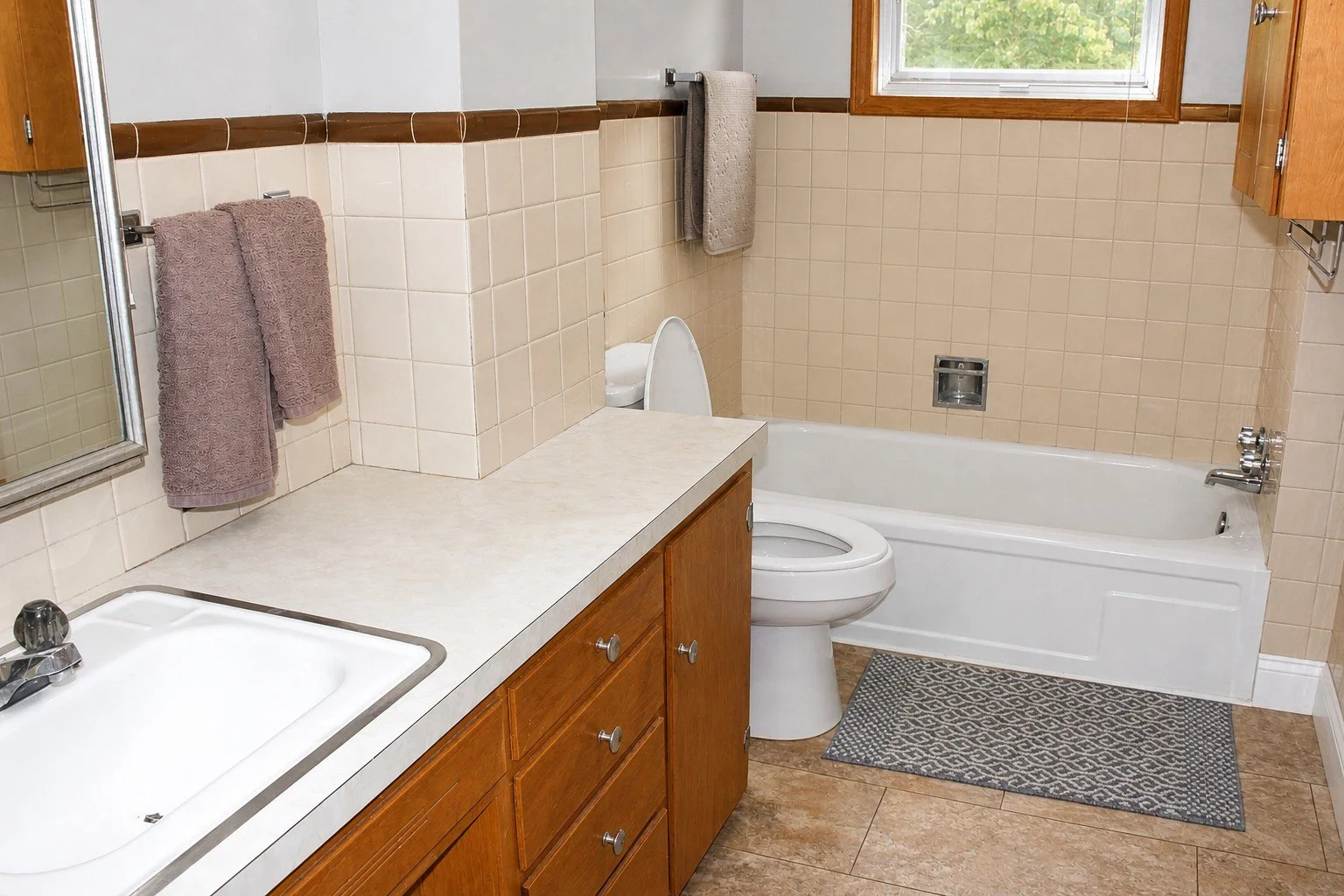 Bathroom with a beige tiled wall, a white bathtub, a toilet, a window, dark towels on a wall hook, and a beige countertop with wooden cabinets.