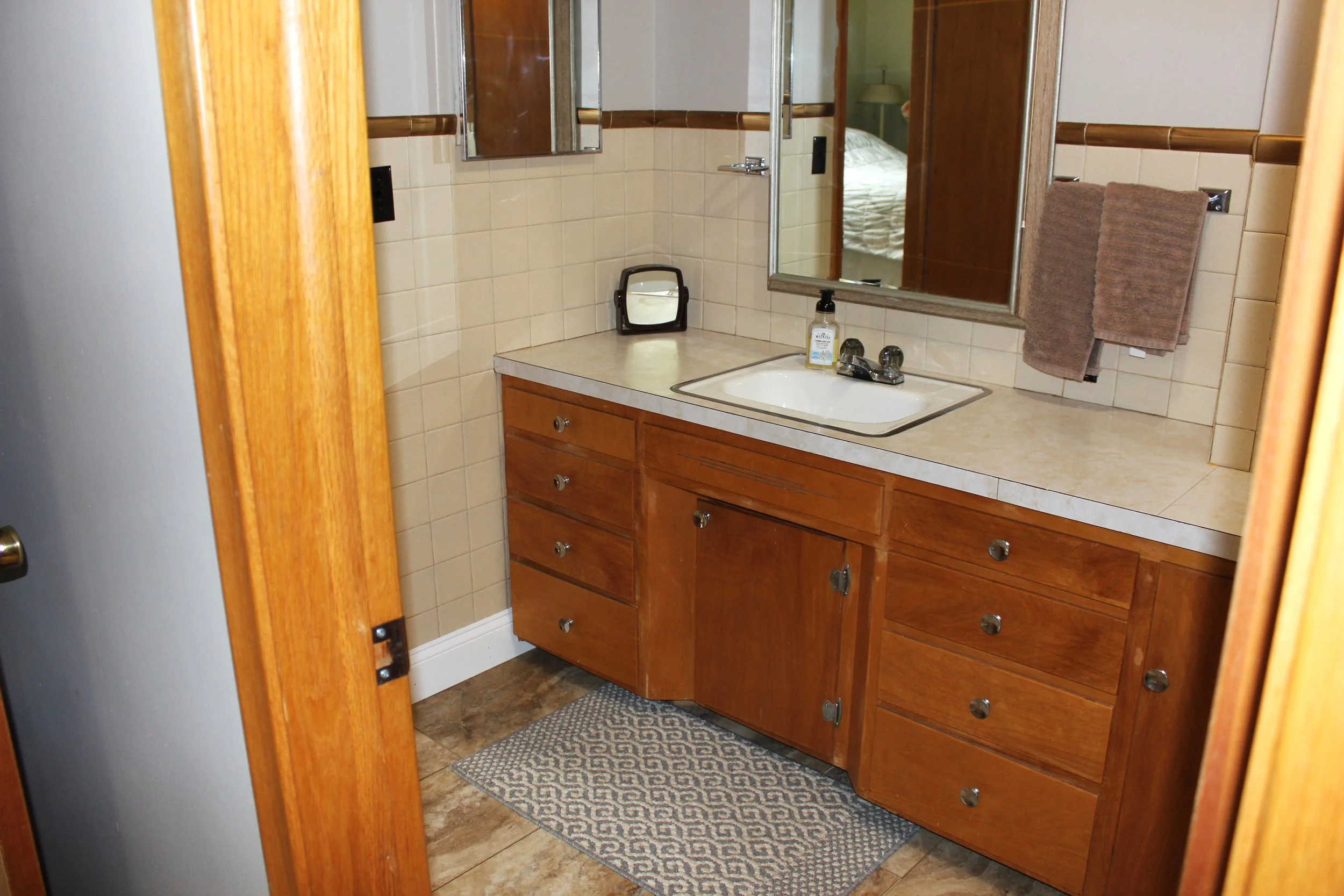A bathroom with a wooden vanity and a beige countertop, a sink, a mirror, and beige tiled walls. There are two brown towels hanging on the wall.
