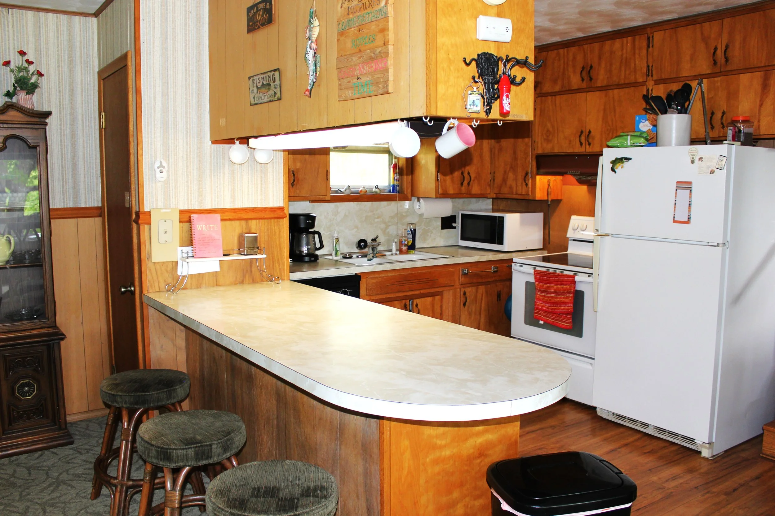 A cozy kitchen with wooden cabinets, a white refrigerator, microwave, coffee maker, and a counter with barstools. Various decorations and kitchen utensils are visible.
