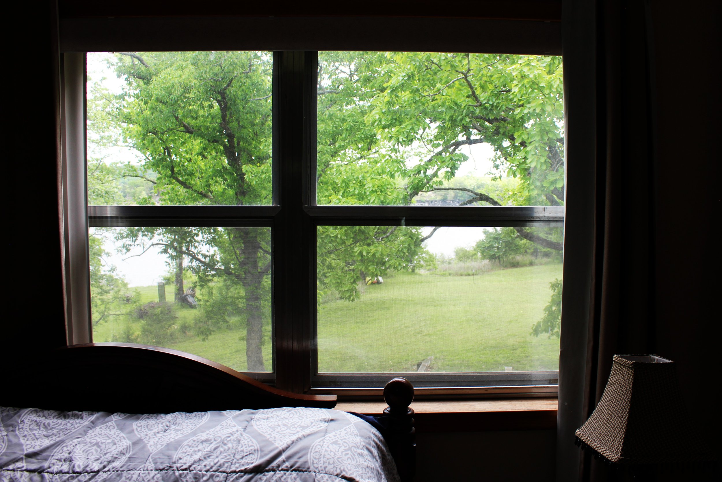 View through a window showing green trees and a grassy yard outside. Inside, part of a bed with patterned white and gray bedding and a wooden headboard is visible, along with a woven chair on the right.