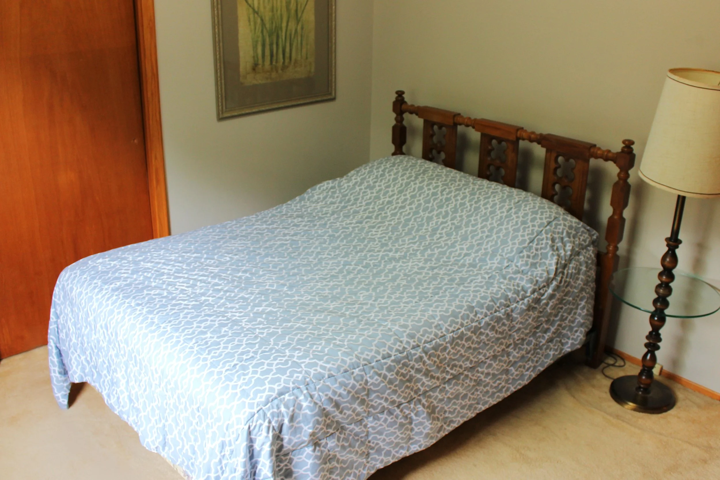 A wooden bed with a patterned blue and white bedspread in a bedroom. Next to the bed is a glass-topped nightstand with a tall, beige lampshade. On the wall above the bed hangs a framed floral picture. There is a wooden wardrobe on the left side of th