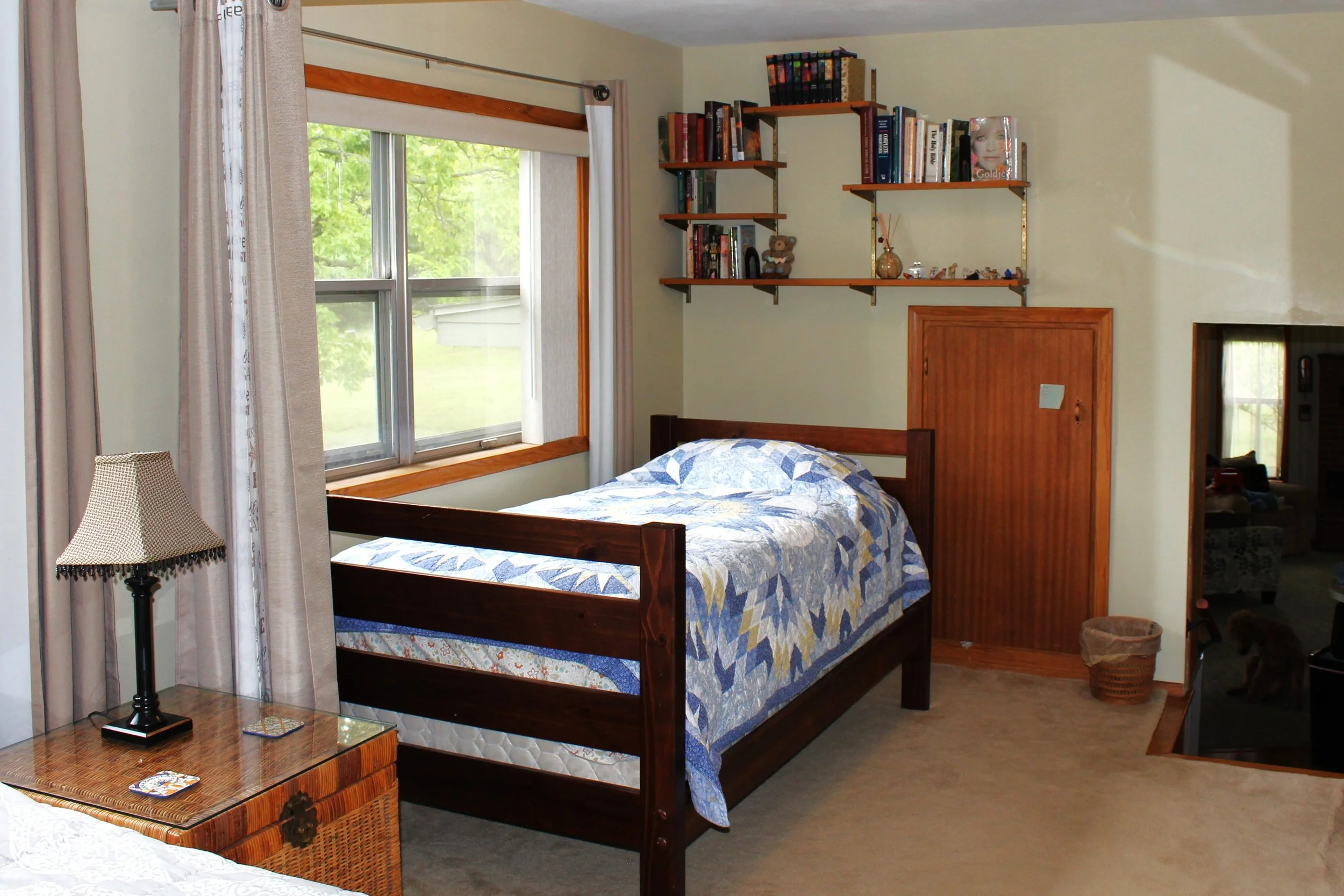 A bedroom with a wooden bed frame, a quilted bedspread with blue, yellow, and white patterns, a nearby wicker basket, a table lamp with a dotted lampshade, a window with beige curtains showing green trees outside, and a wall-mounted wooden shelf with