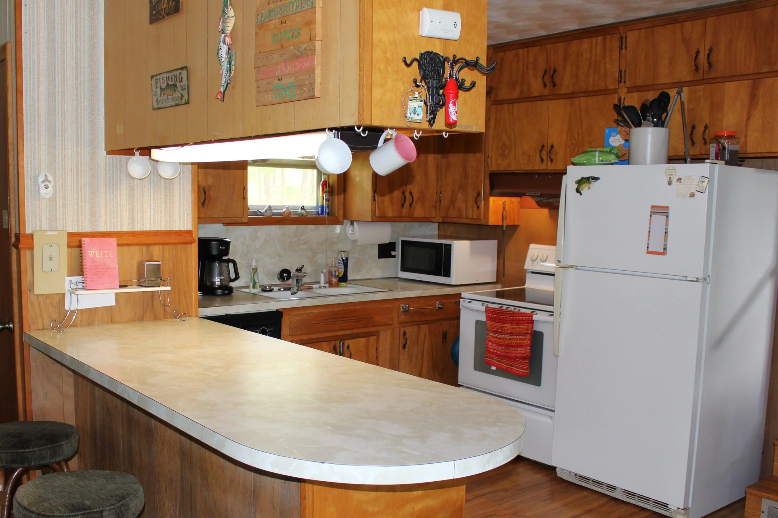 A cozy kitchen with wooden cabinets, a white refrigerator, a microwave, and various kitchen utensils and decor items. There is a marble-like countertop and a small window above the sink. The walls are covered with wood paneling and some wall hangings