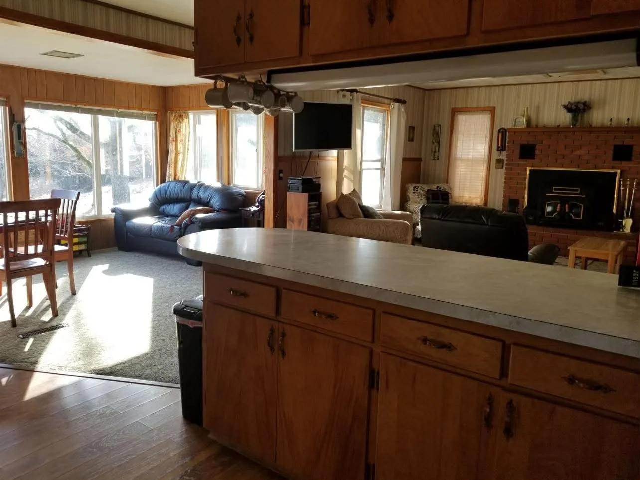 Living room with large windows, blue sofa, beige armchair, black recliner, brick fireplace, TV, and wooden furniture, viewed from the kitchen counter.