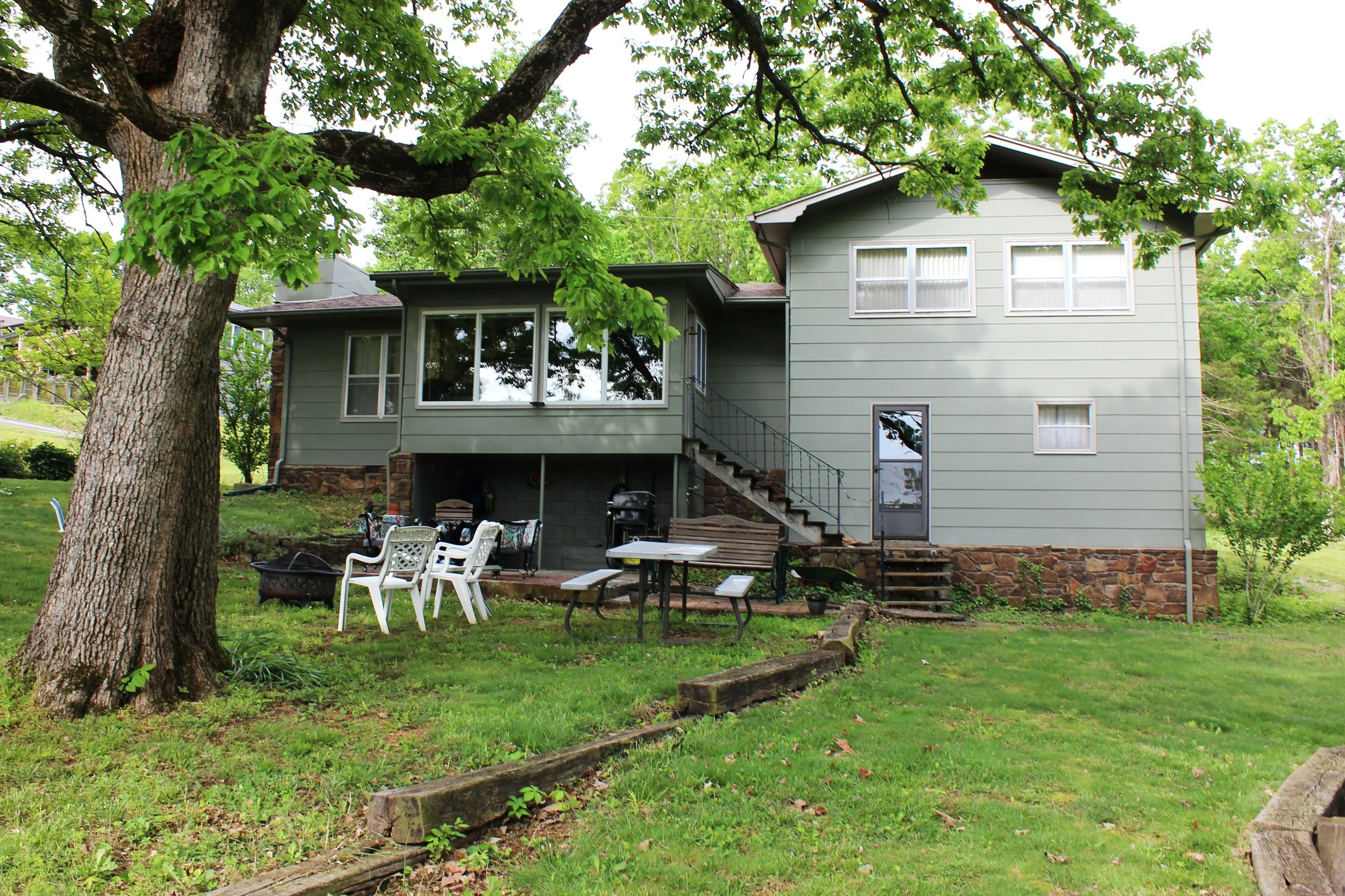 Backyard scene with a two-story house, large tree in the foreground, and outdoor furniture including chairs and a table on a grassy lawn.