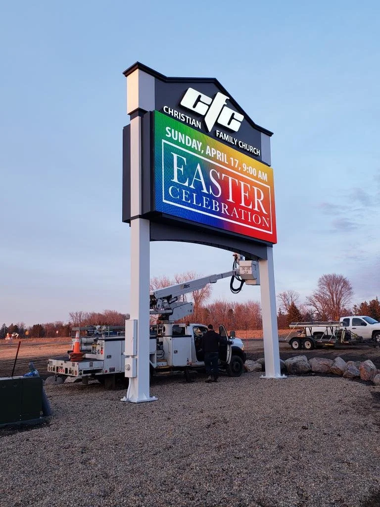 A large church sign with the name 'Christian Family Church' and an announcement for Easter celebration on April 17 at 9:00 AM. A worker is using a lift to install or repair the sign, which is on a gravel lot with trees and a few parked vehicles in the background.