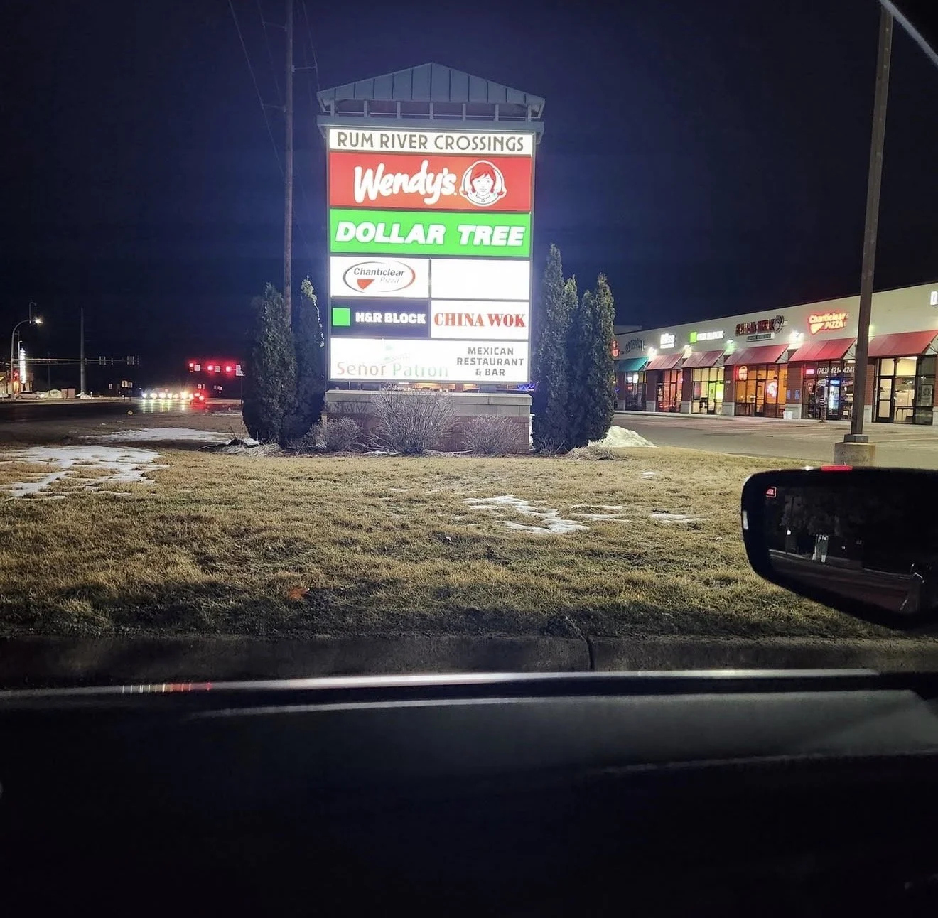 A brightly lit shopping center sign at night displays store names including Wendy's, Dollar Tree, Chanticleer, H & R Block, China Wok, and Señor Patron Mexican Restaurant & Bar. In the background, there are storefronts with illuminated signs and a pa