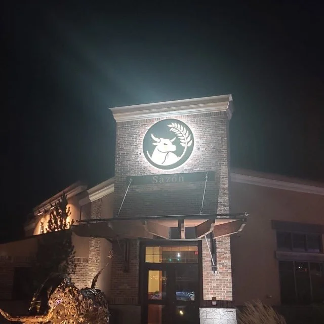 The exterior of a restaurant at night with a lit deer logo and the word "Sazon" on the storefront.