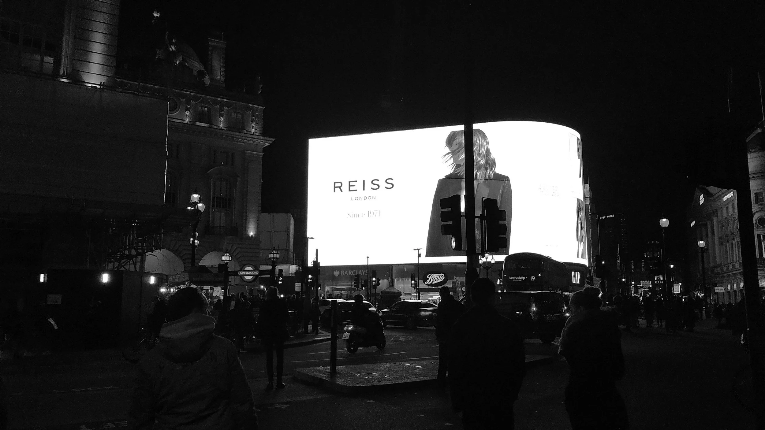 Nighttime scene of Piccadilly Circus in London with a large illuminated digital advertisement screen showing the Reiss logo and an image of a woman from the back. Silhouettes of people and vehicles are visible in the foreground.