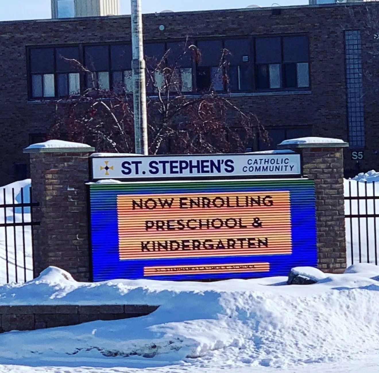 A sign outside St. Stephen's Catholic Community announces the enrollment of preschool and kindergarten classes, with snow on the ground and a building in the background.