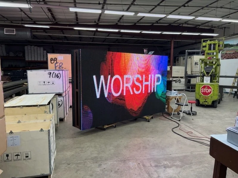 Large digital screen displaying the word 'WORSHIP' in bright, colorful text inside a warehouse or industrial space, with equipment and boxes around.