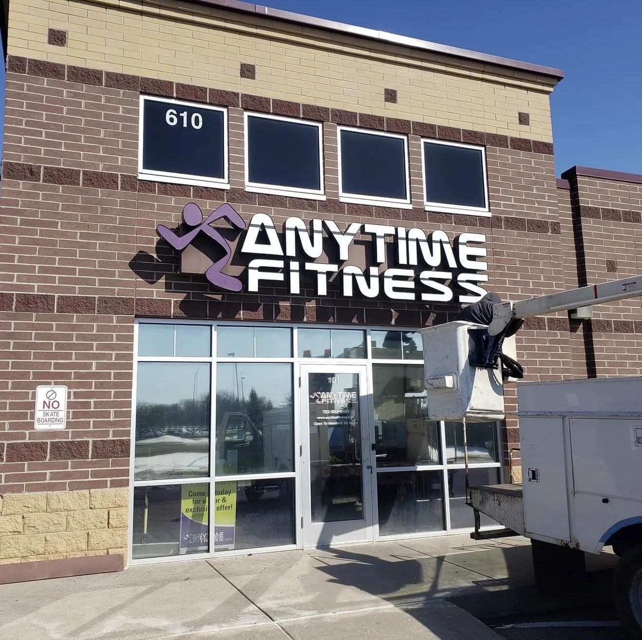 A fitness center called Anytime Fitness with a purple and white sign on a brick building. There is a no skateboarding sign on the wall, and a utility lift truck is parked outside.