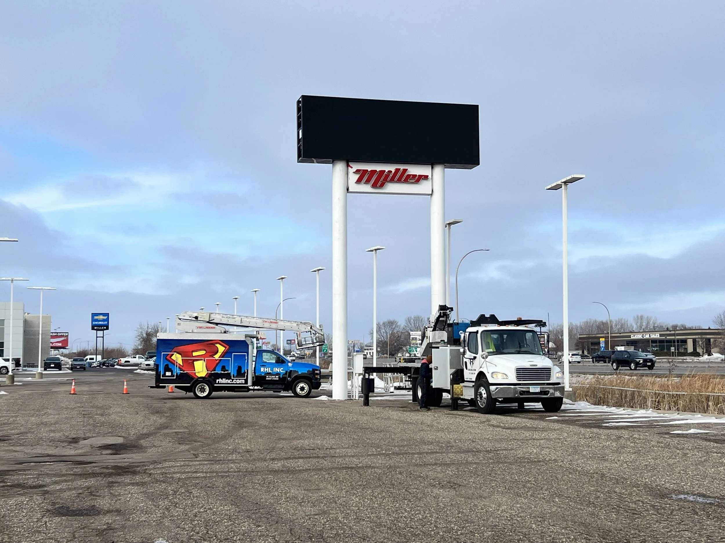 A parking lot with utility trucks, one with a Superman logo, near a Miller's sign, and a large empty digital billboard.