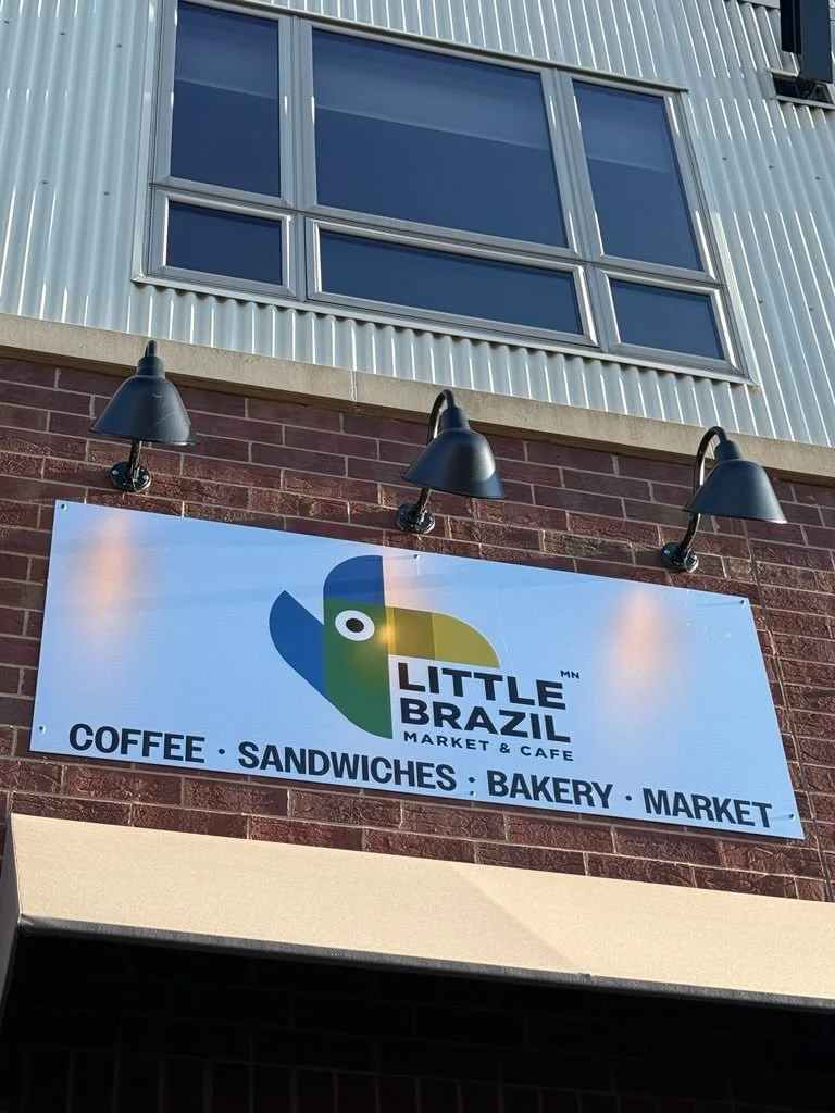 Exterior signage for Little Brazil Market & Cafe, featuring a colorful parrot logo. The sign indicates the establishment offers coffee, sandwiches, bakery items, and market goods.