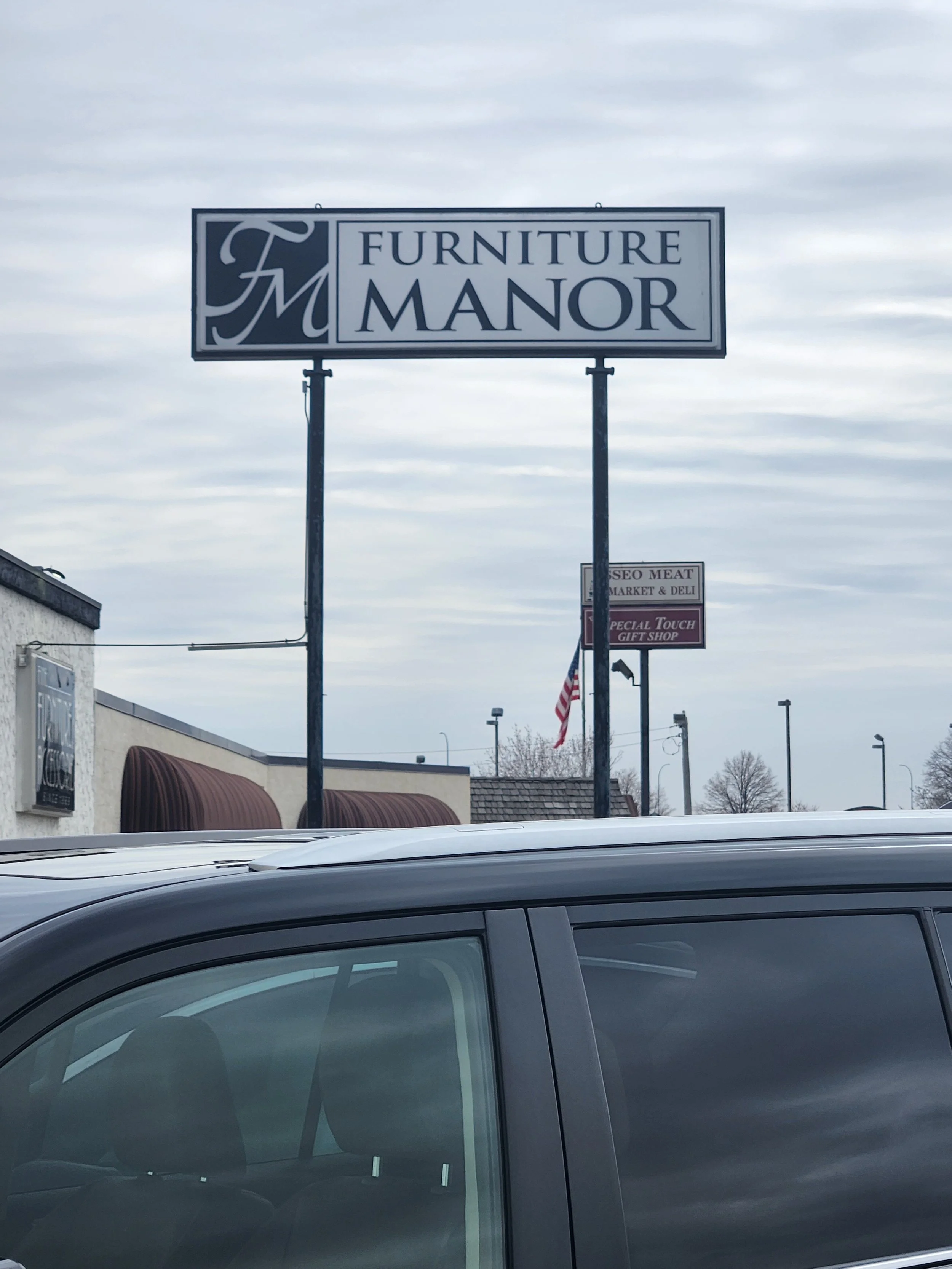 Sign for Furniture Manor store in a parking lot with cloudy sky.