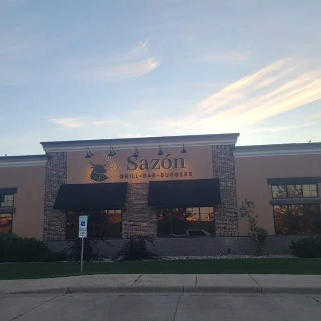 Exterior of Sazon restaurant with sign displaying grill, bar, and burgers, and an accessible parking sign in front.