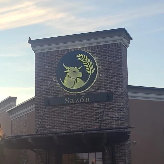 The exterior of a building with a sign featuring a reindeer holding a wheat stalk logo and the word "Sazón" beneath it.