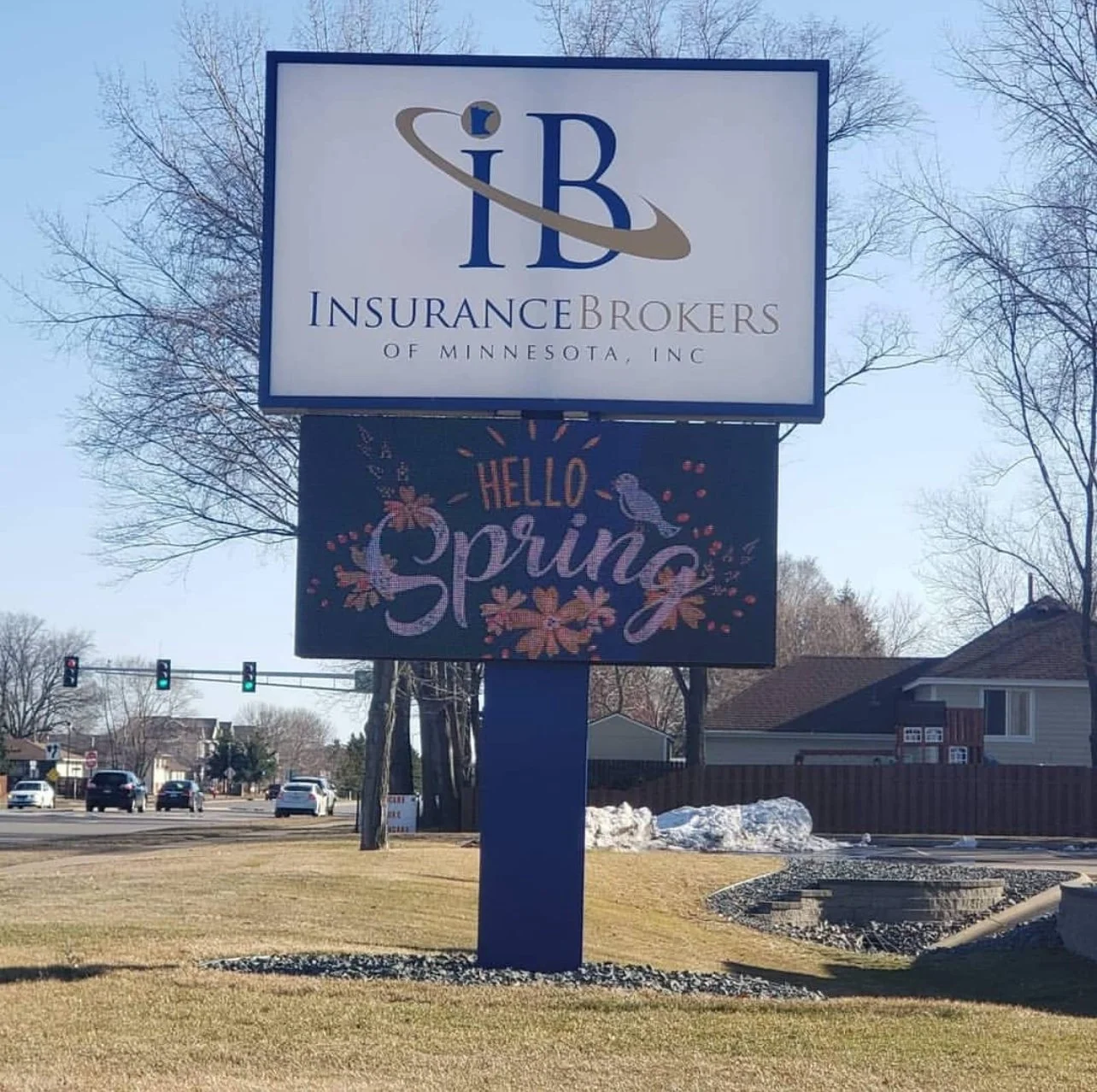 A billboard display in front of a residential neighborhood with a blue sky. The top sign reads 'Insurance Brokers of Minnesota, Inc' with a logo featuring a large 'IB' and a curved swoosh. Below it, another sign says 'Hello Spring' with floral and bi