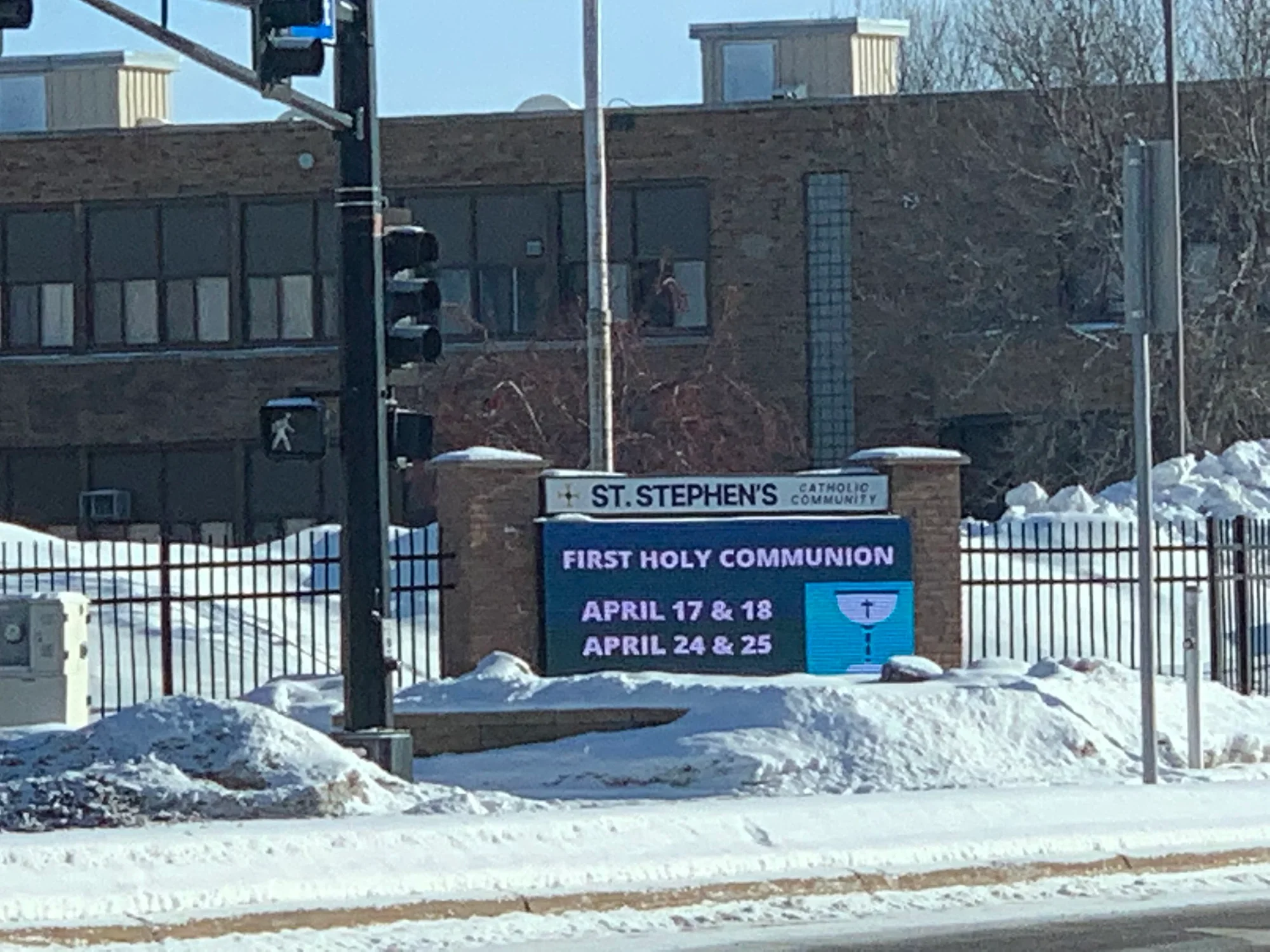 A street scene with snow on the ground, showing a sign for St. Stephen's Catholic Community announcing First Holy Communion dates for April 17 & 18 and April 24 & 25. There are traffic lights, a fence, a pole, and a building in the background.