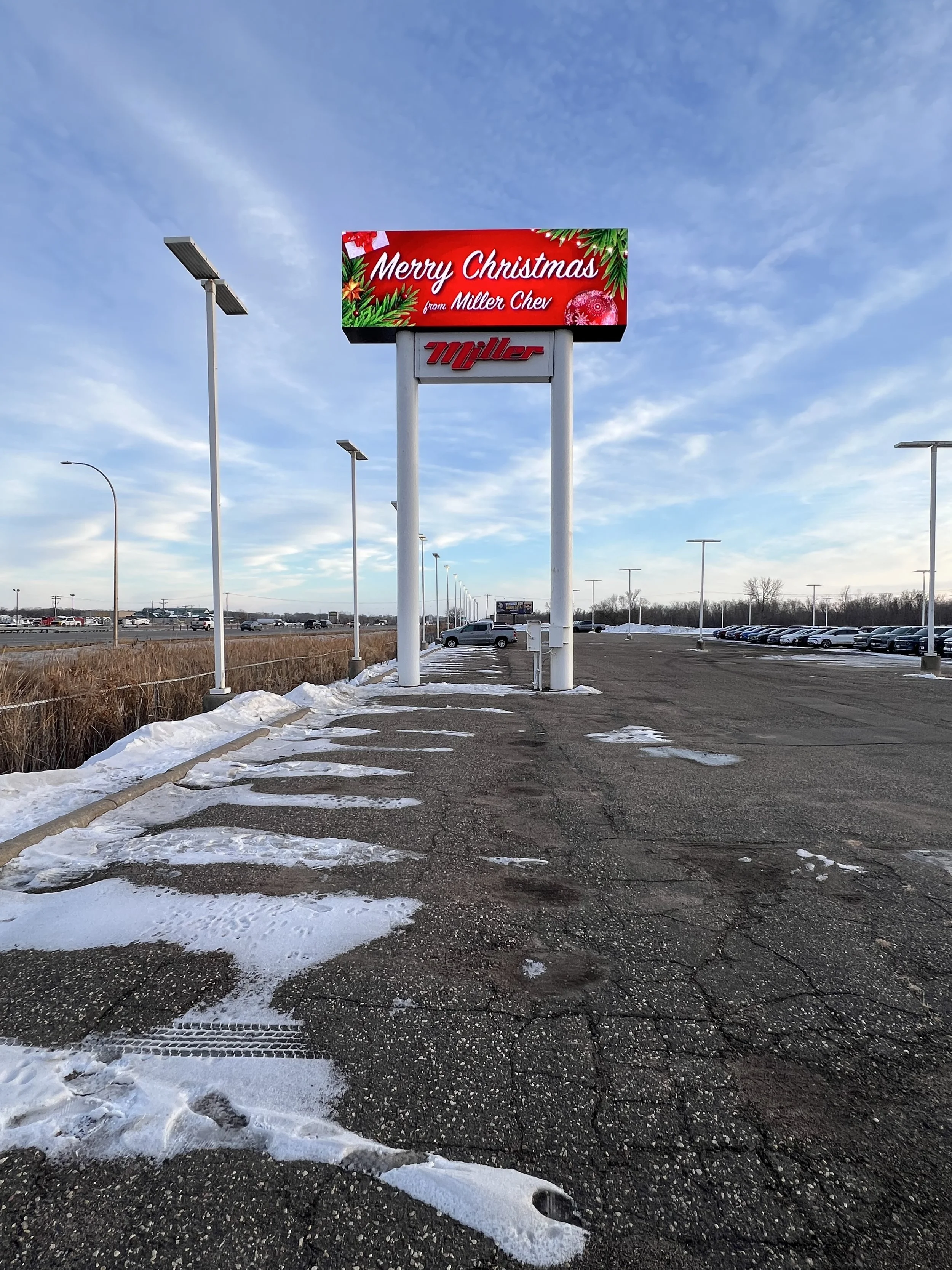 Empty parking lot with scattered snow patches, street lamps, and a large sign displaying holiday greeting 'Merry Christmas from Miller Cher' over a bright blue sky.