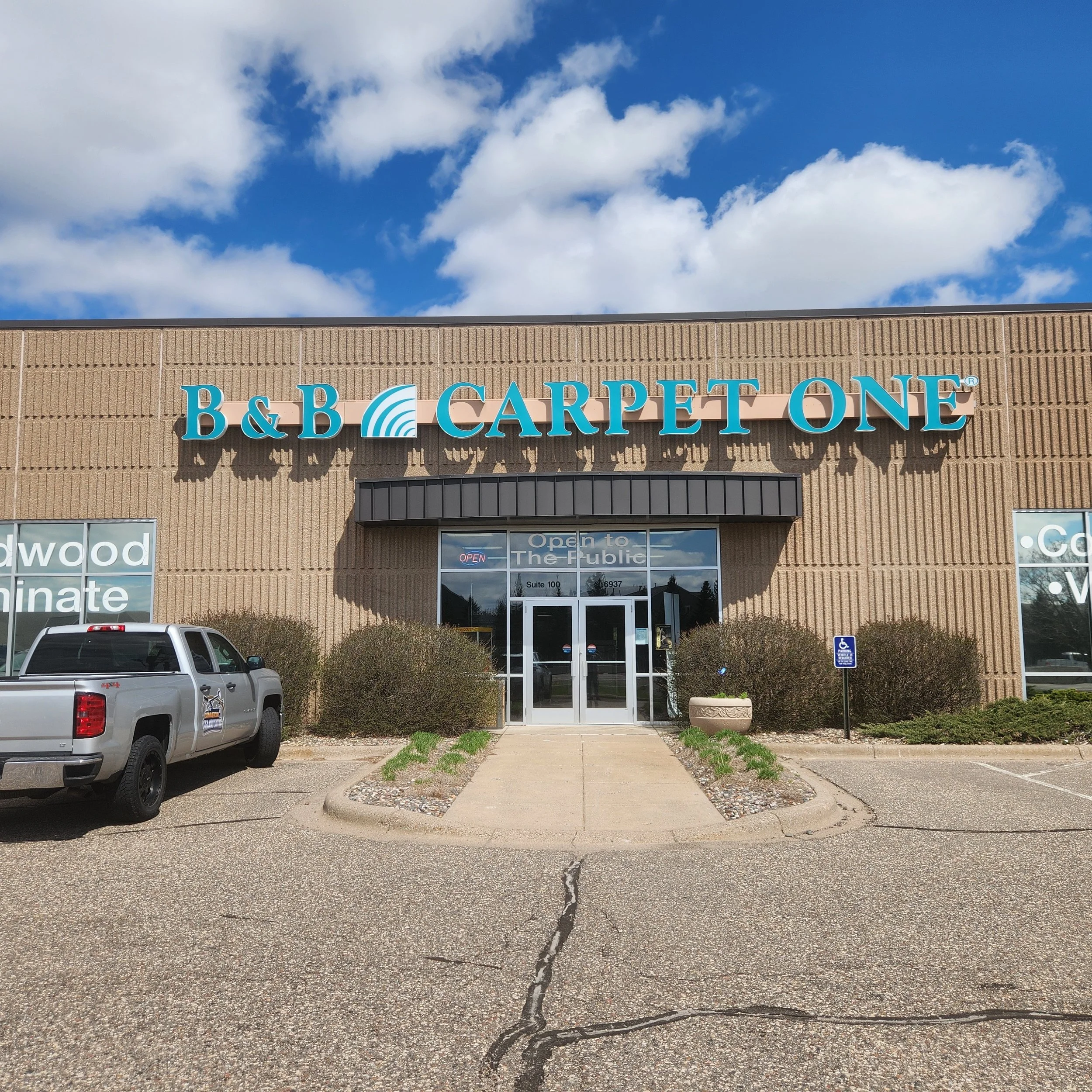 Front view of a building with a sign reading 'B&B Carpet One'. There is a parking lot with a silver pickup truck and a handicap parking space. The building has glass doors and windows, with shrubbery and planters surrounding the entrance, under a par