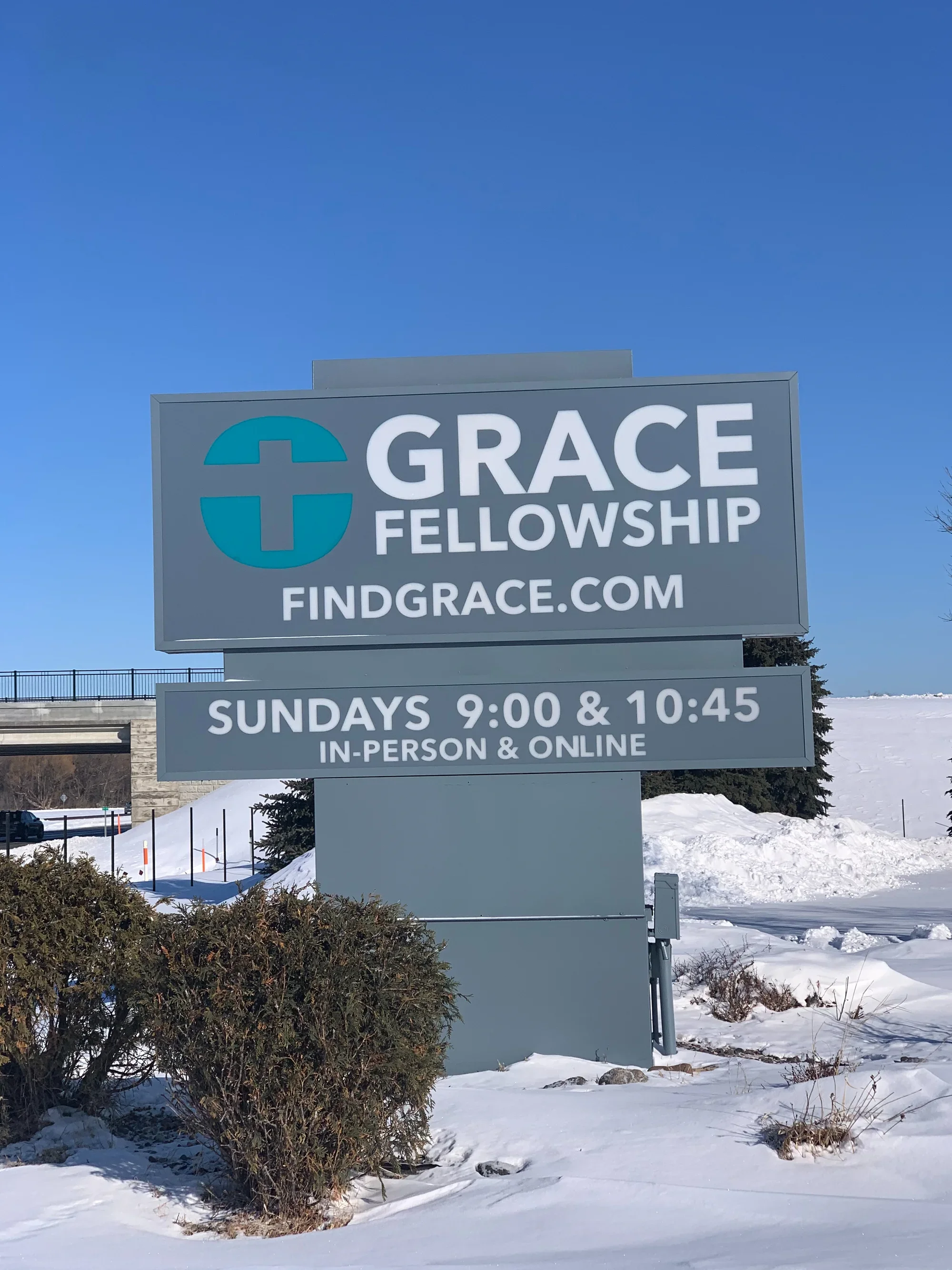 Sign for Grace Fellowship with service times on a snowy day, displaying Sundays at 9:00 and 10:45, in-person and online, with clear blue sky in the background.