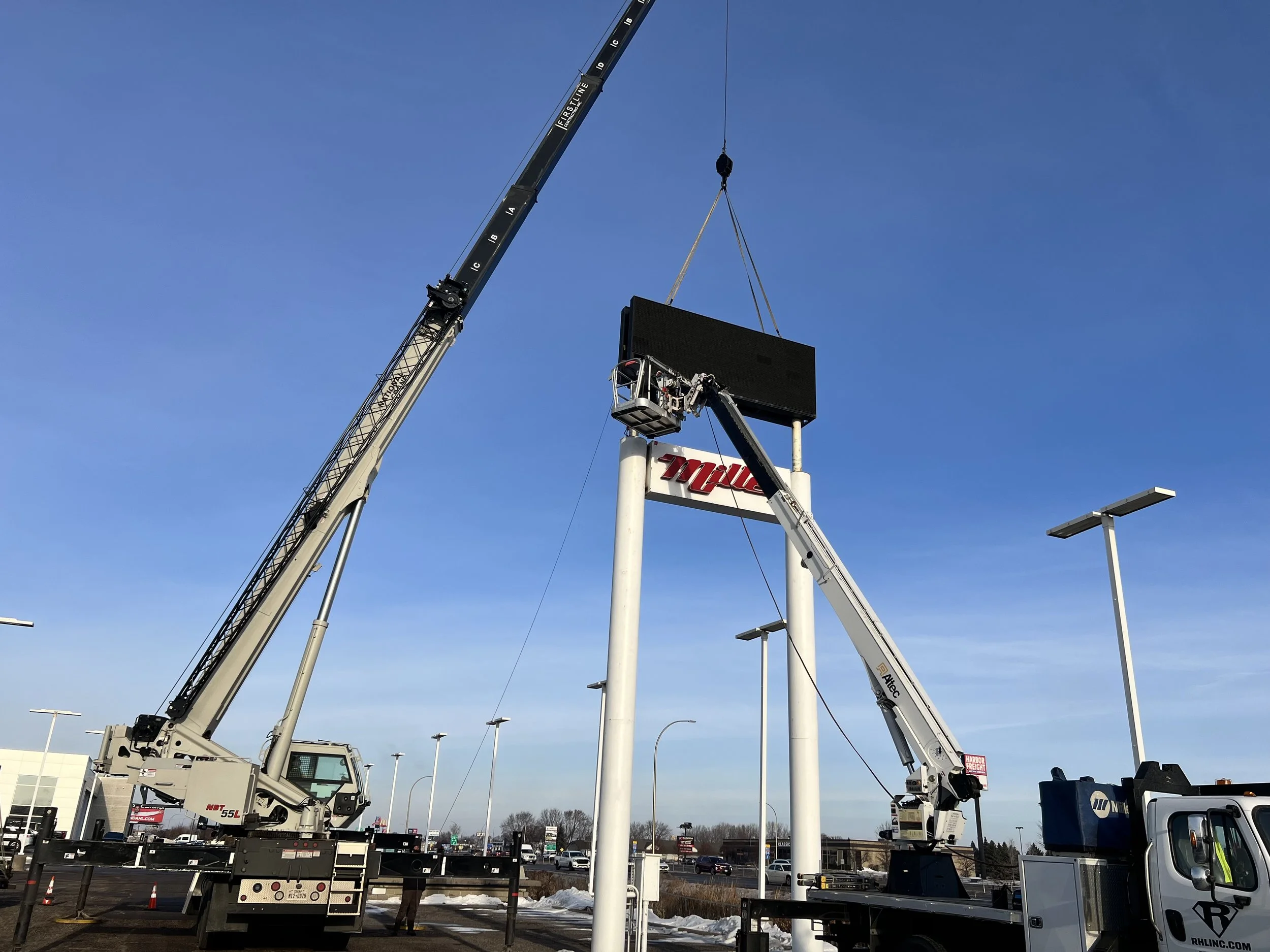 Two cranes are installing or maintaining a large digital billboard at a retail store parking lot on a clear day. The billboard is mounted on tall white support poles, and part of a store sign is visible beneath it.