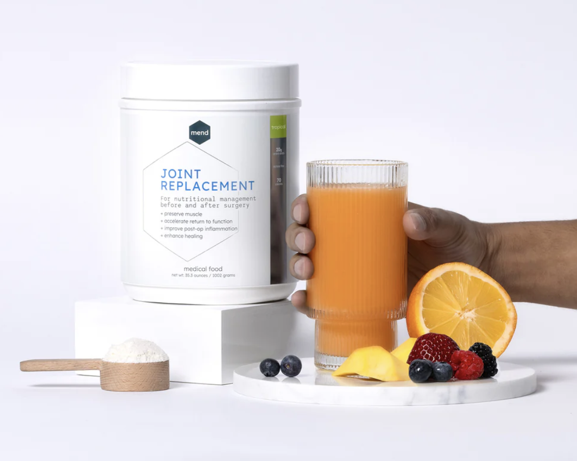 A tub of joint replacement nutritional supplement beside a glass of orange juice, with sliced orange, blueberries, raspberries, blackberries, and a lemon wedge in front.