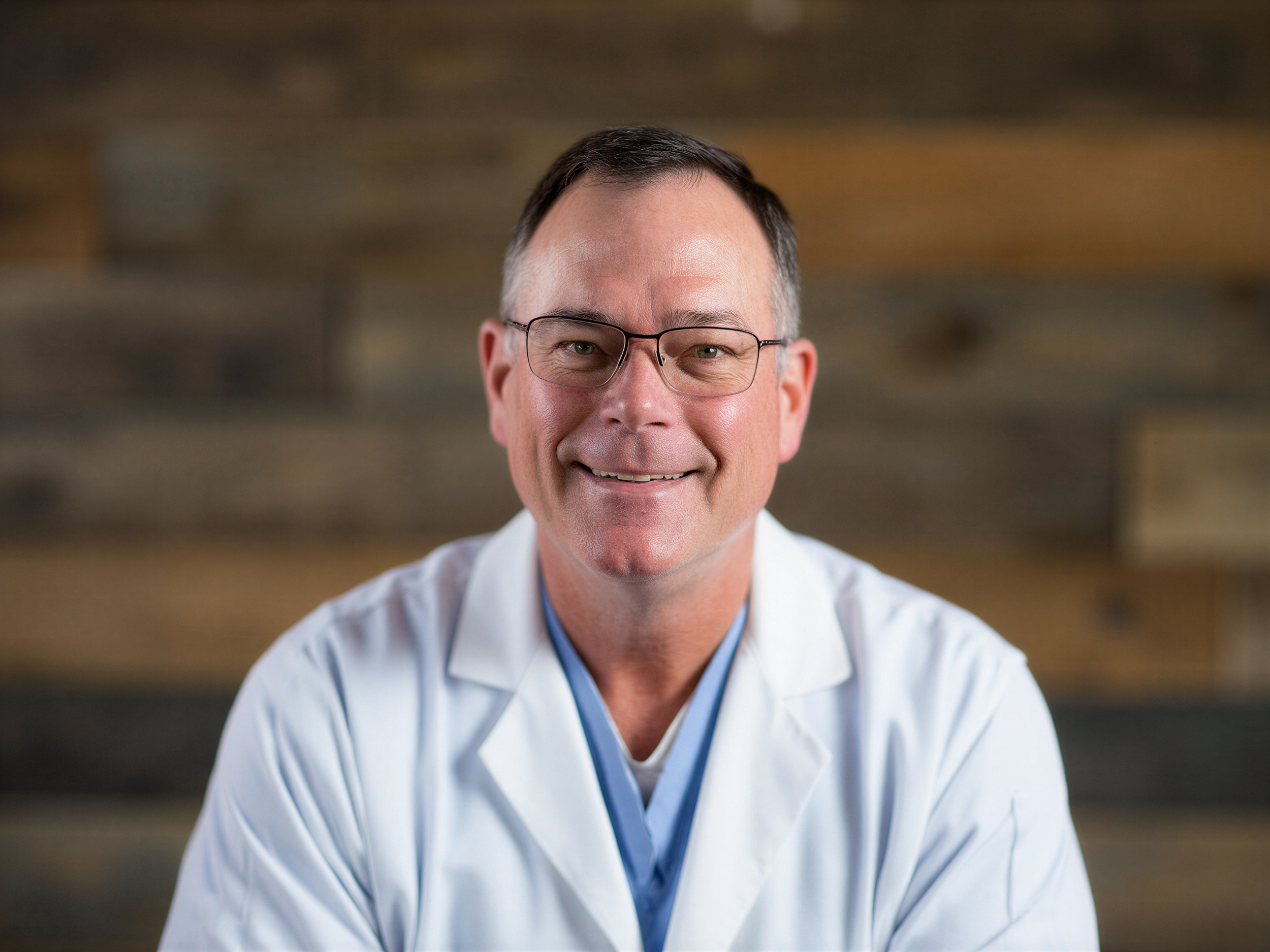 Portrait of a smiling middle-aged man wearing glasses and a white coat, with a wooden wall background.