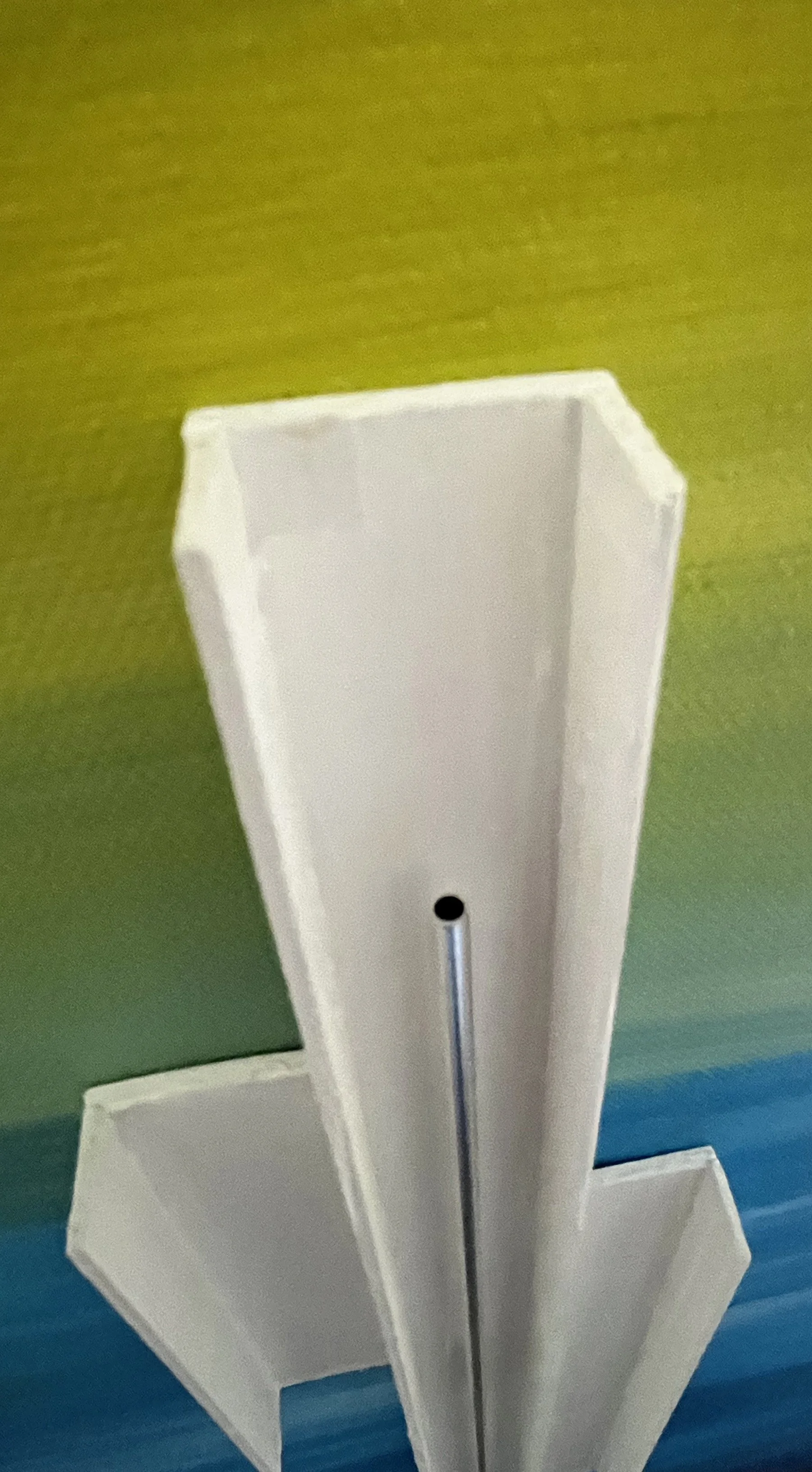 Carrie Caulkins - Close-up of a white geometric sculpture with a metal rod against a background of green, yellow, and blue colors.