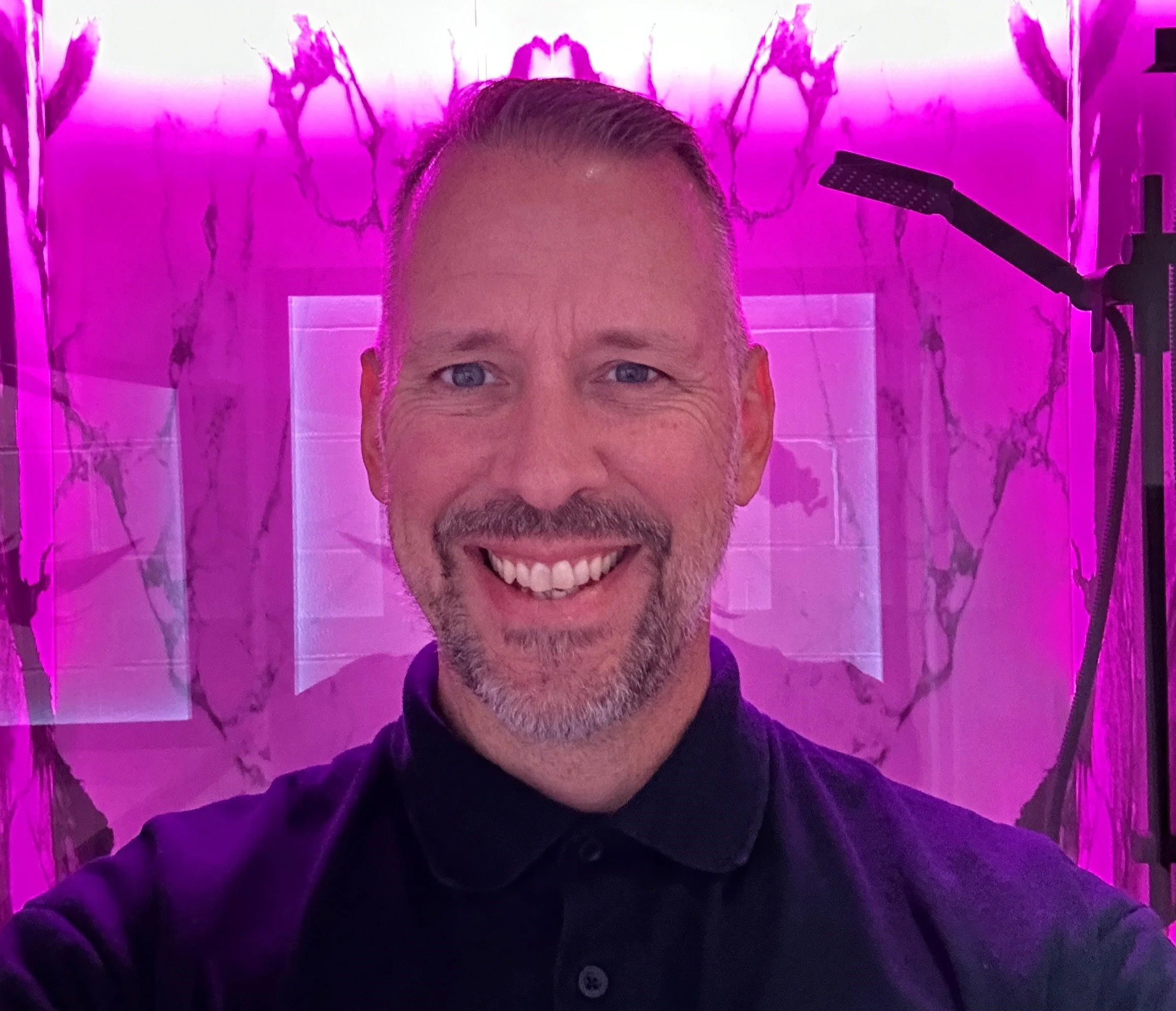 A smiling man with short, gray hair and a beard taking a selfie in front of a pink-lit marble-patterned wall with a showerhead on the right.