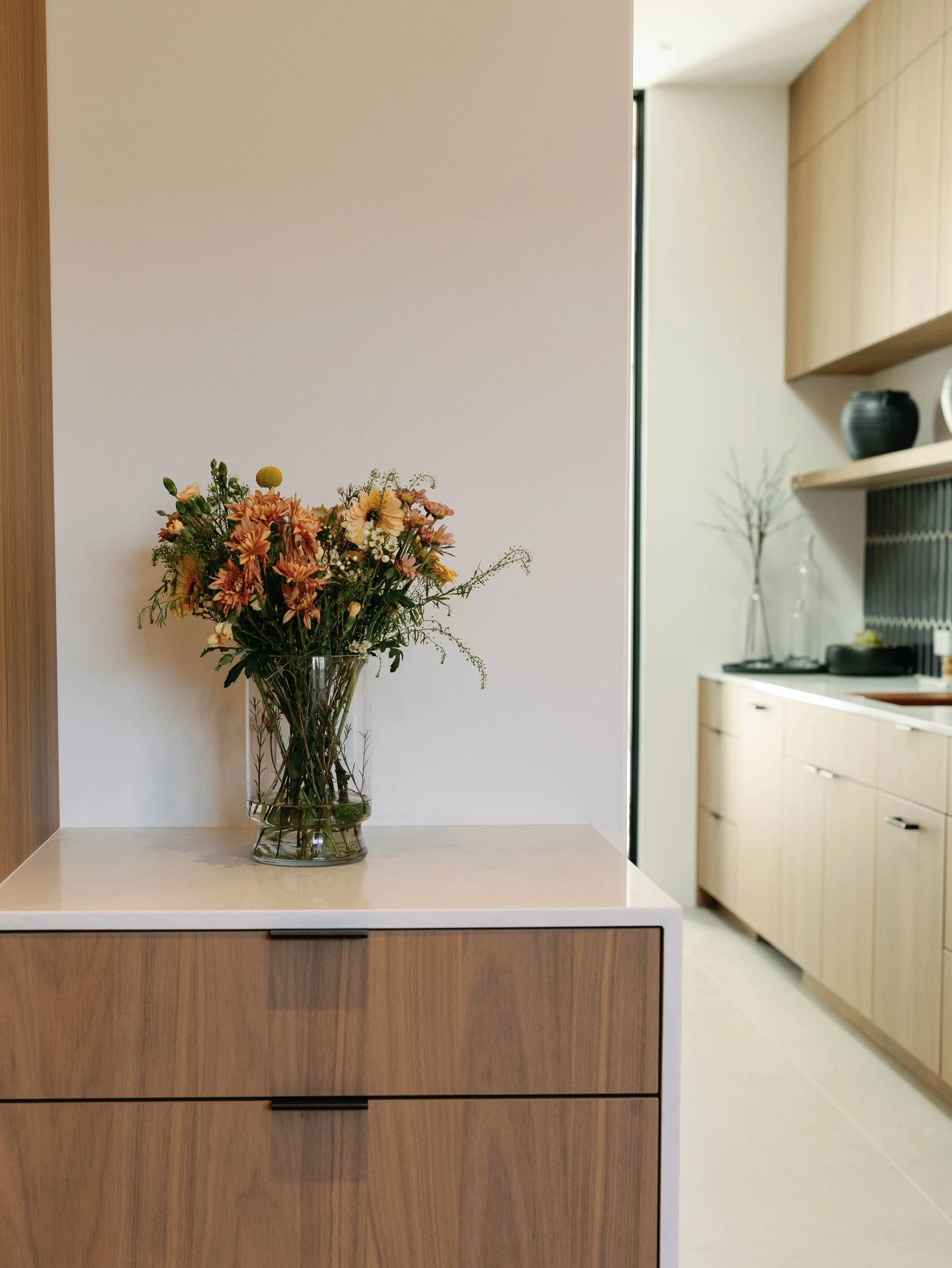 A glass vase with a bouquet of colorful flowers on a wooden cabinet in a modern, minimalist kitchen.