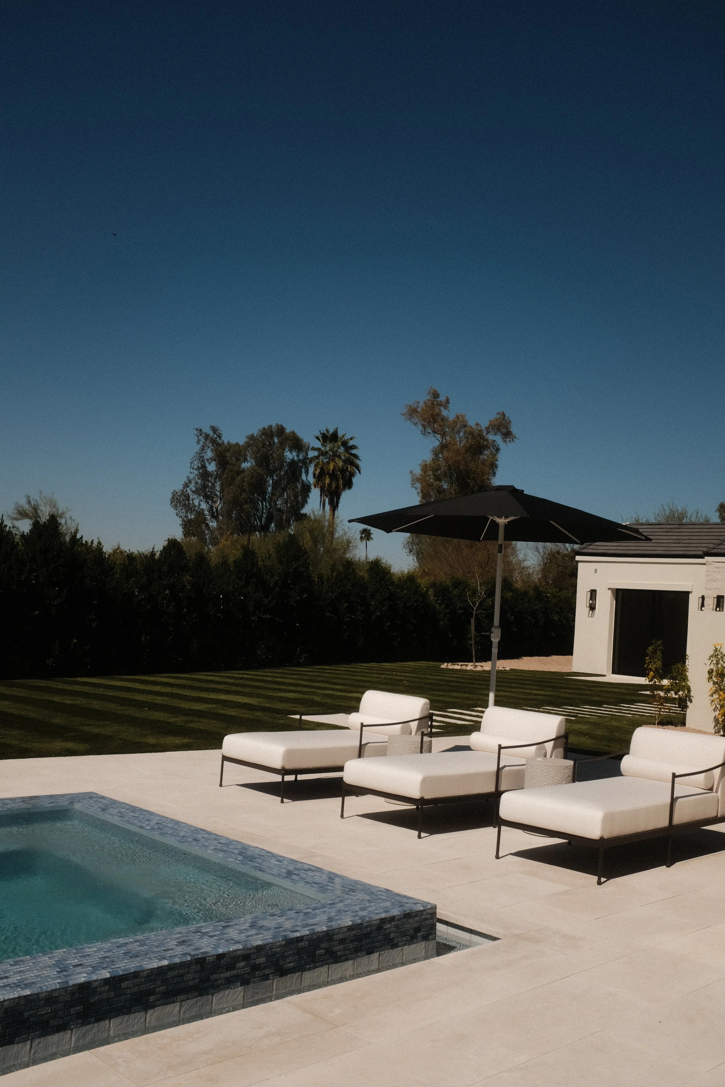 A backyard with a swimming pool, white lounge chairs, a black umbrella, a grassy yard, trees, and a small white building under a blue sky.