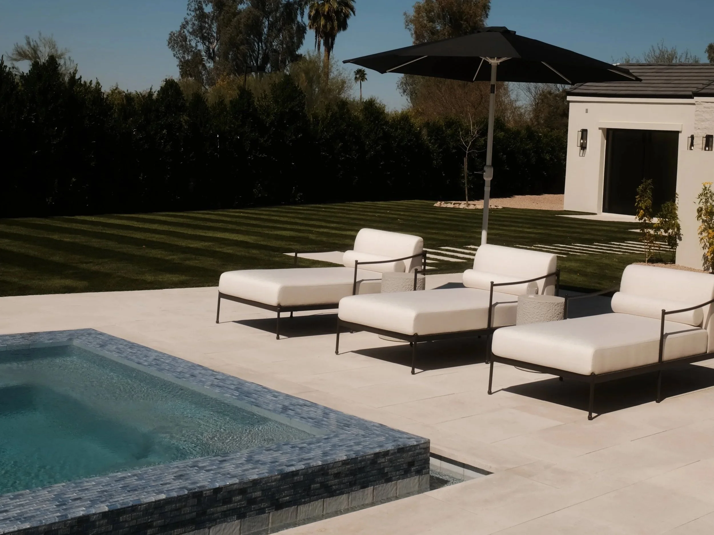 Luxury backyard with a swimming pool, white outdoor lounge chairs, a large black umbrella, a manicured lawn, and a modern house in the background.