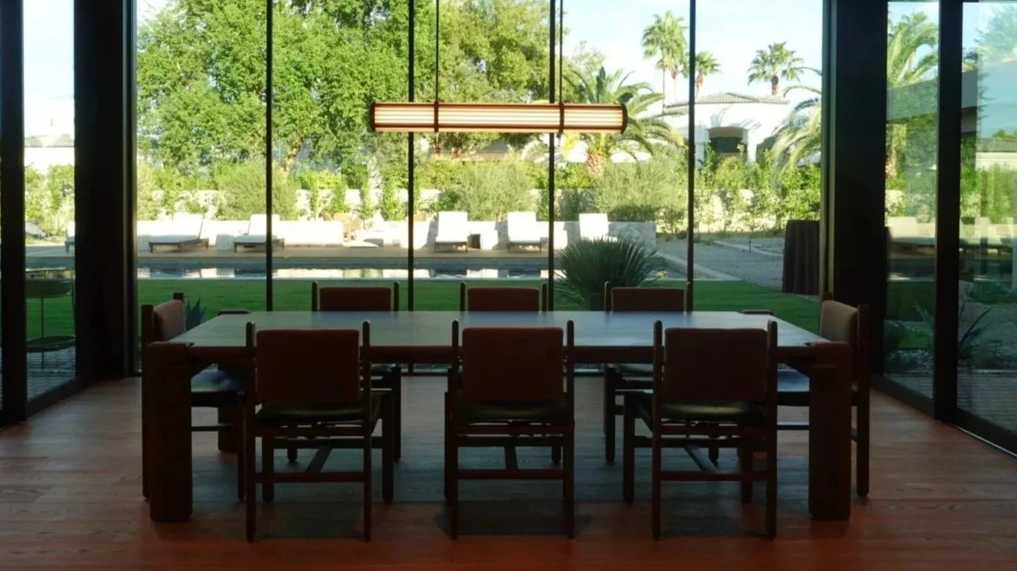 A modern dining room with a large wooden table and eight chairs, situated in front of floor-to-ceiling glass windows that overlook a pool, palm trees, and lush greenery outside, with a hanging rectangular light fixture above the table.