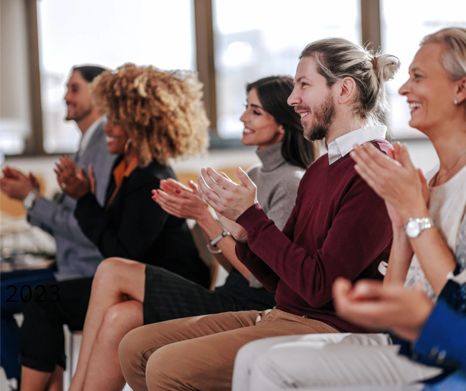 A group of diverse people sitting and clapping together, smiling at an indoor event.