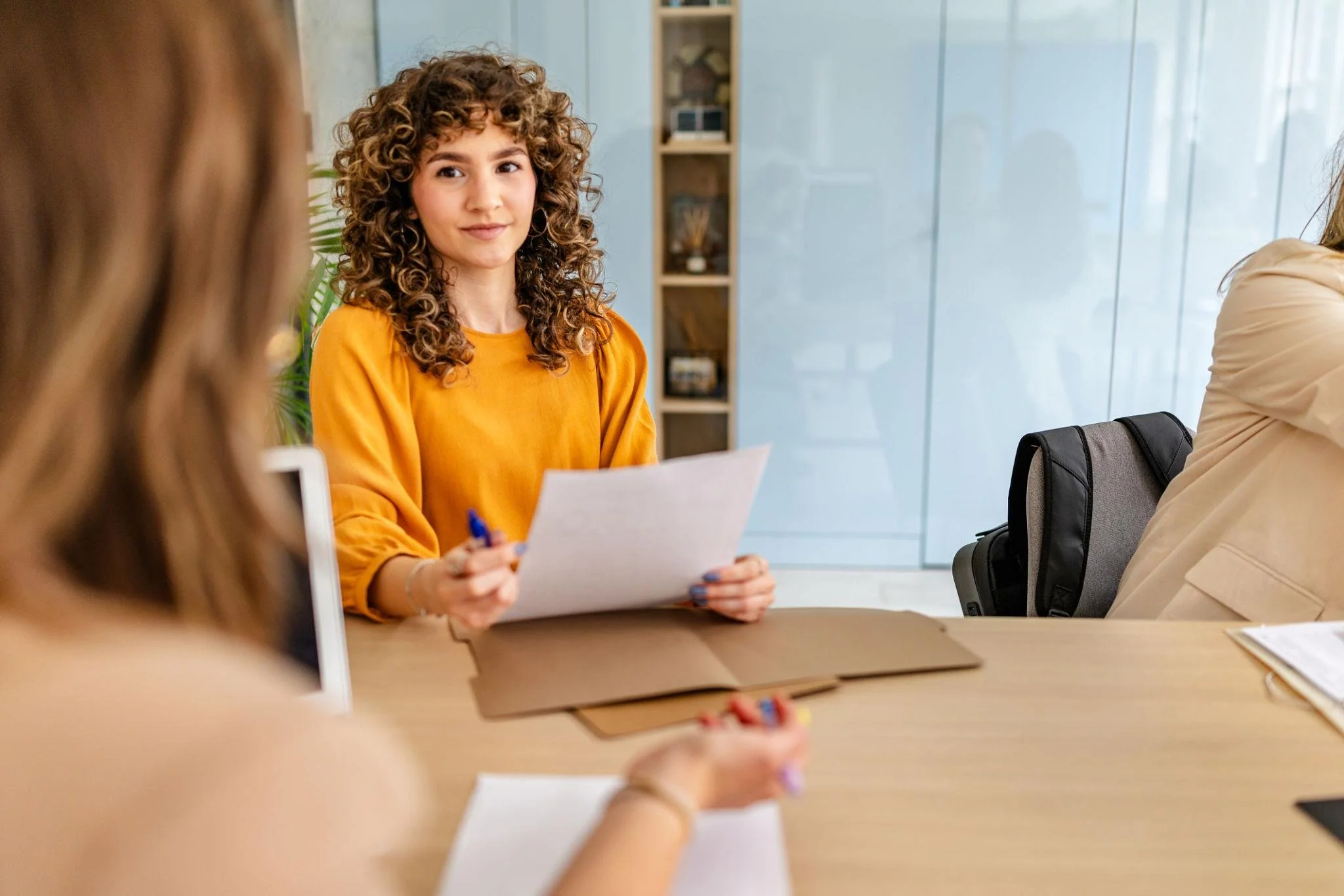 A woman with curly hair in a yellow shirt sitting at a table holding a piece of paper, engaged in conversation with others during a meeting in an office.