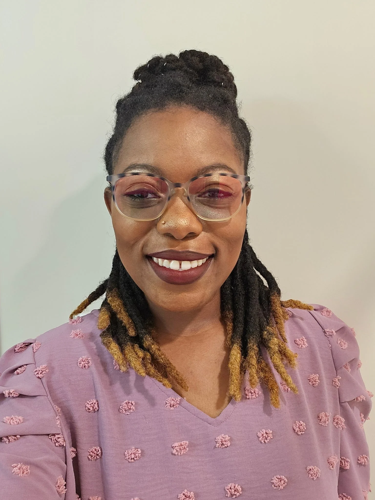 A woman smiling, wearing glasses and a purple top with textured floral patterns.