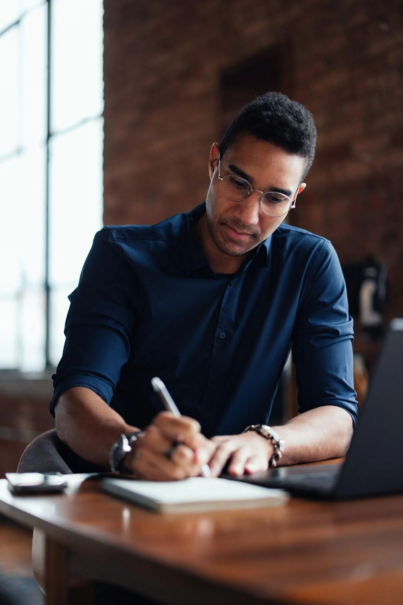 A young man with glasses, wearing a navy blue shirt, sitting at a desk writing in a notebook with a pen, next to a laptop in a room with brick walls and large windows.