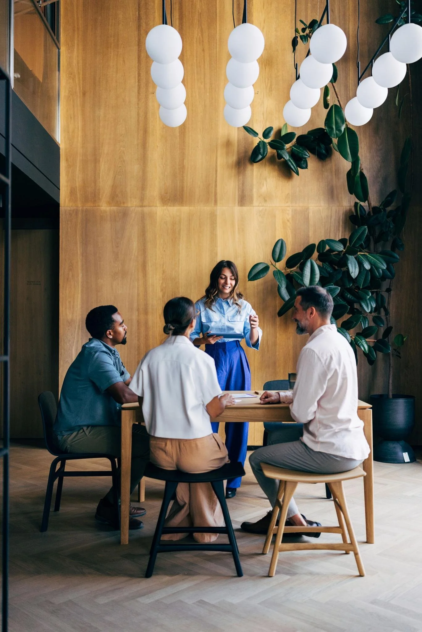 A woman standing and presenting at a table with three seated people inside a modern office with wood panel walls, hanging white spherical light fixtures, and large green plants.