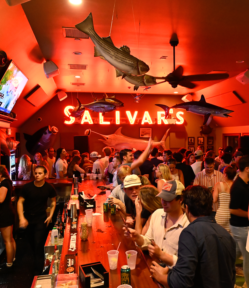 Crowded bar with people socializing, under hanging large fish models, neon sign reading 'Salivar's', and American-themed decorations.