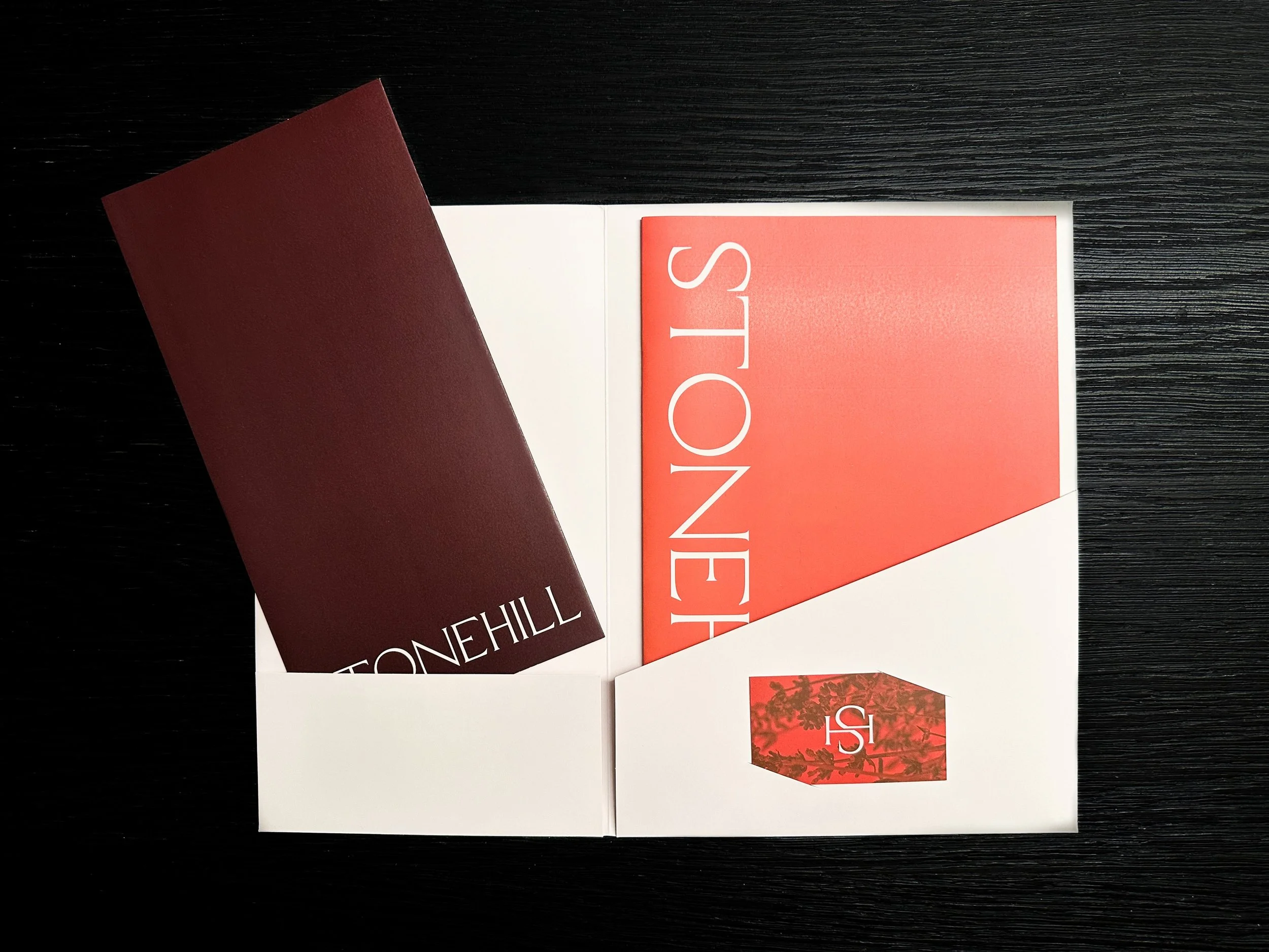 Image of the folder open with maroon floorplan and orange brochure inside on a black wooden surface.