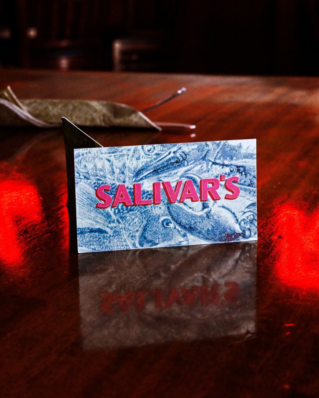 Close-up of a card with the text "SALIVAR'S" in bold pink letters, placed on a reflective wooden table with a blurred background.