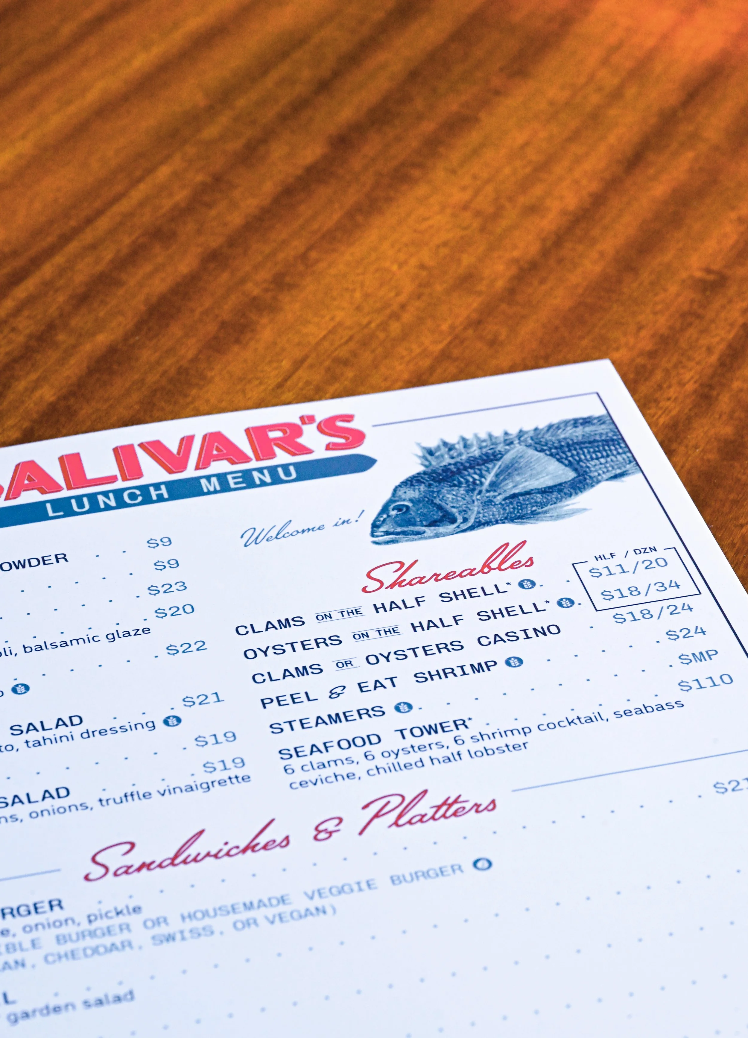 Part of a seafood restaurant menu on a wooden table, with the restaurant name 'Allivars' visible at the top, featuring a fish illustration and various seafood dishes listed.